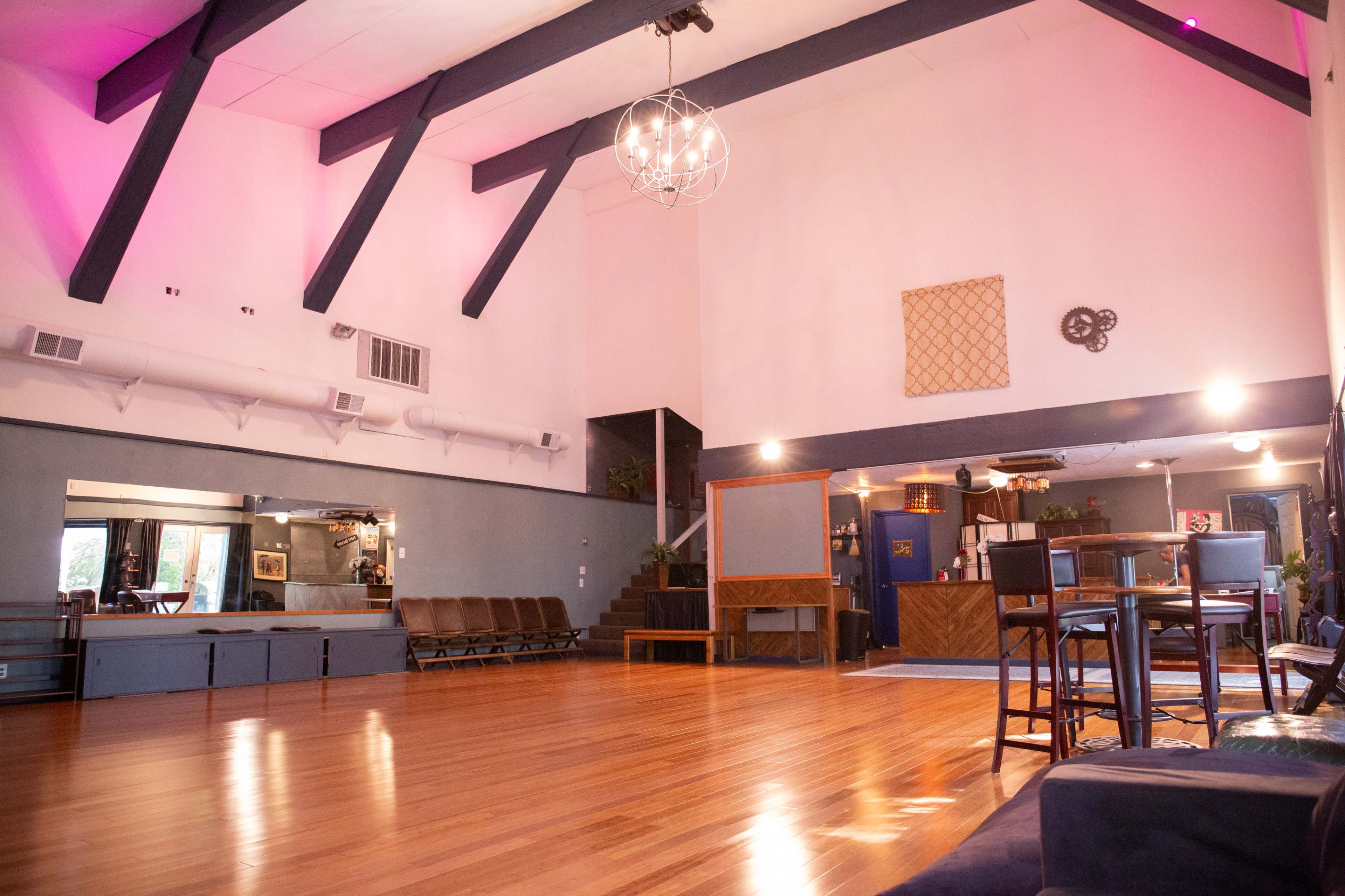A spacious interior with wooden flooring, high ceilings, and a seating area featuring chairs and tables, illuminated by decorative lighting.