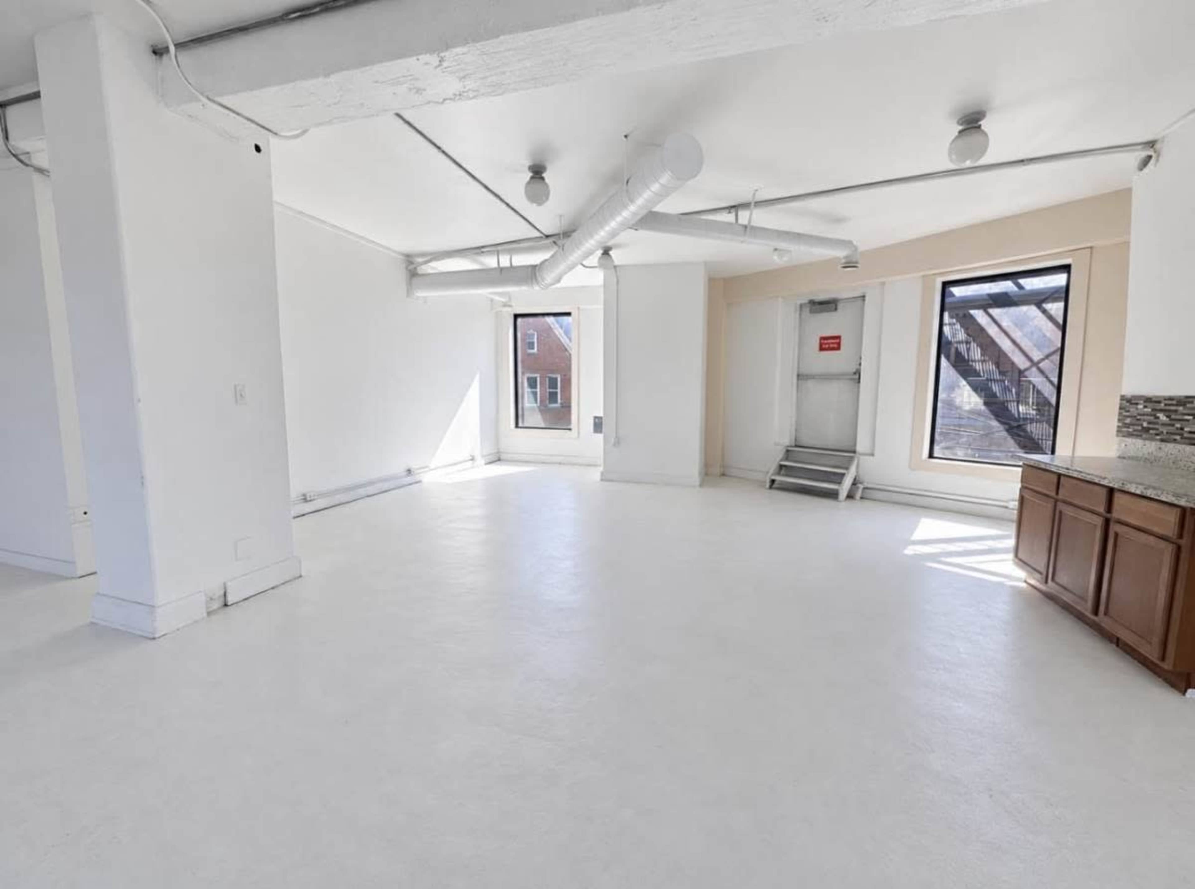 The image shows an empty room with white walls, a light-colored floor, and a large window, along with a small kitchenette area featuring wooden cabinets.