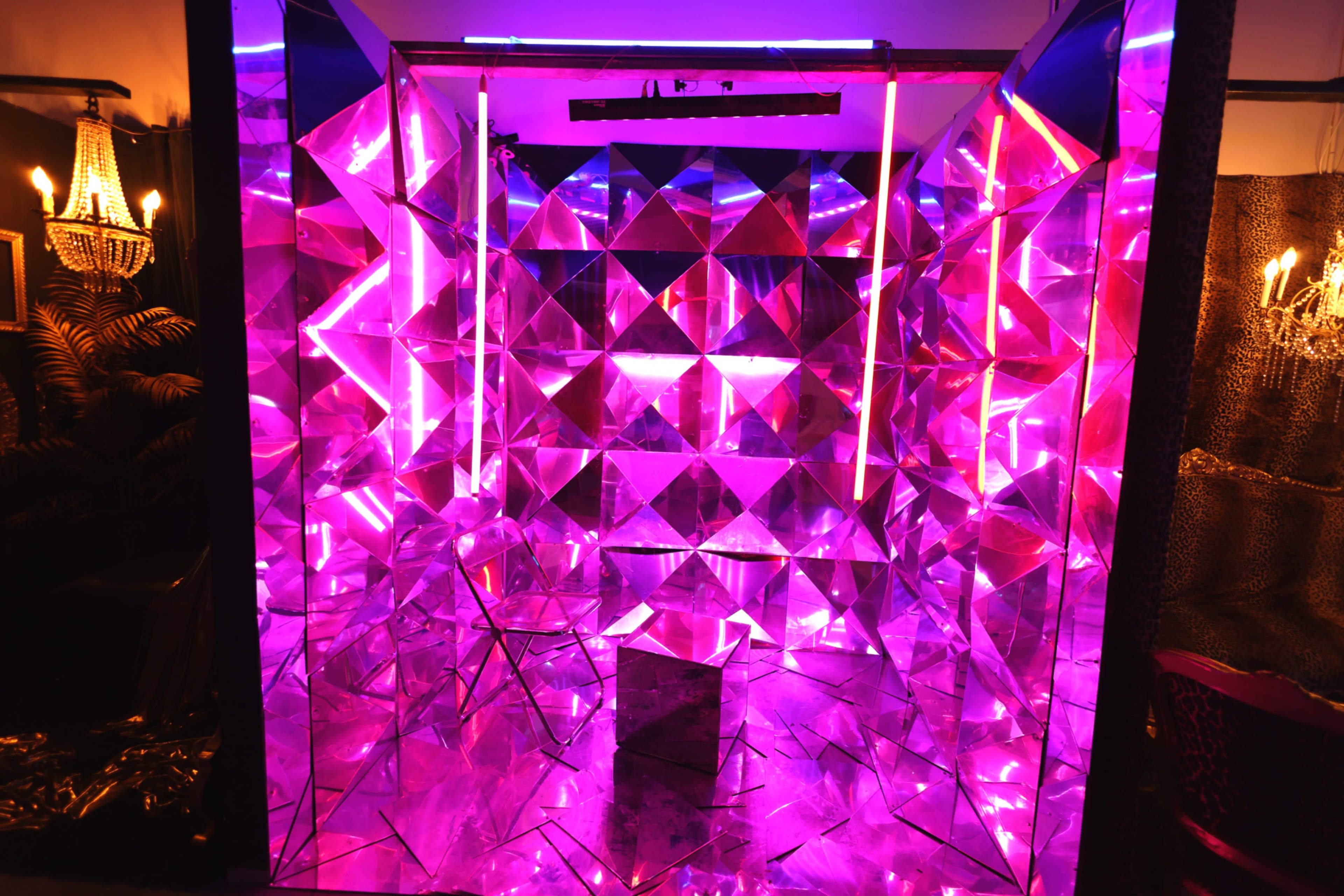 The image shows a mirrored room with geometric panels reflecting vibrant pink and purple lights.
