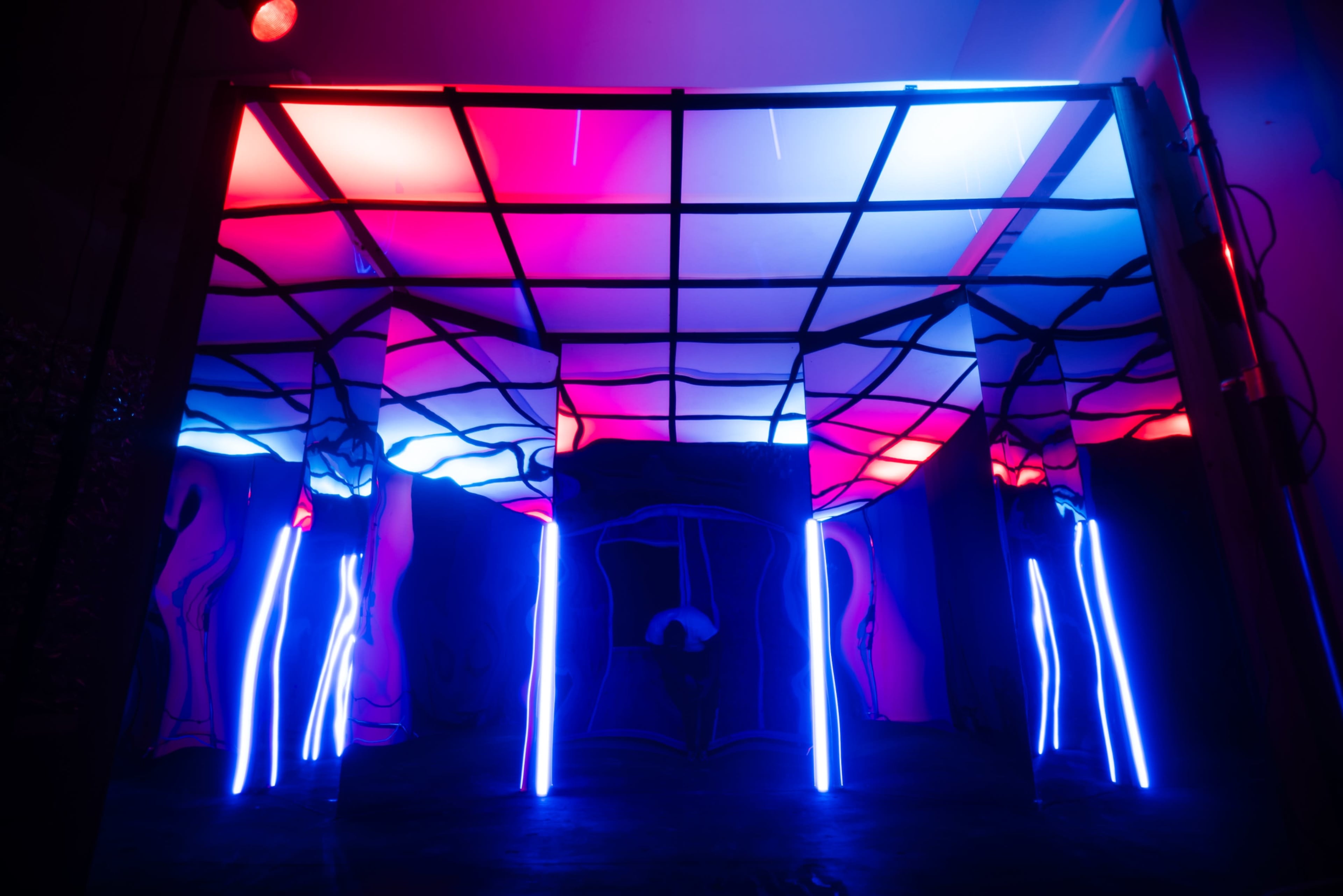 The image shows a dimly lit room with abstract, colorful lighting and a grid-like structure made of illuminated columns.