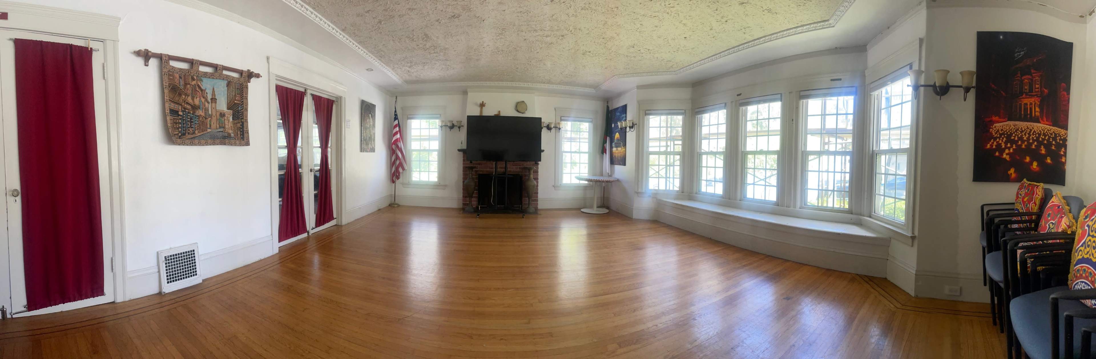 The image shows a panoramic view of a spacious room with wooden floors, large windows, and decorative wall art.