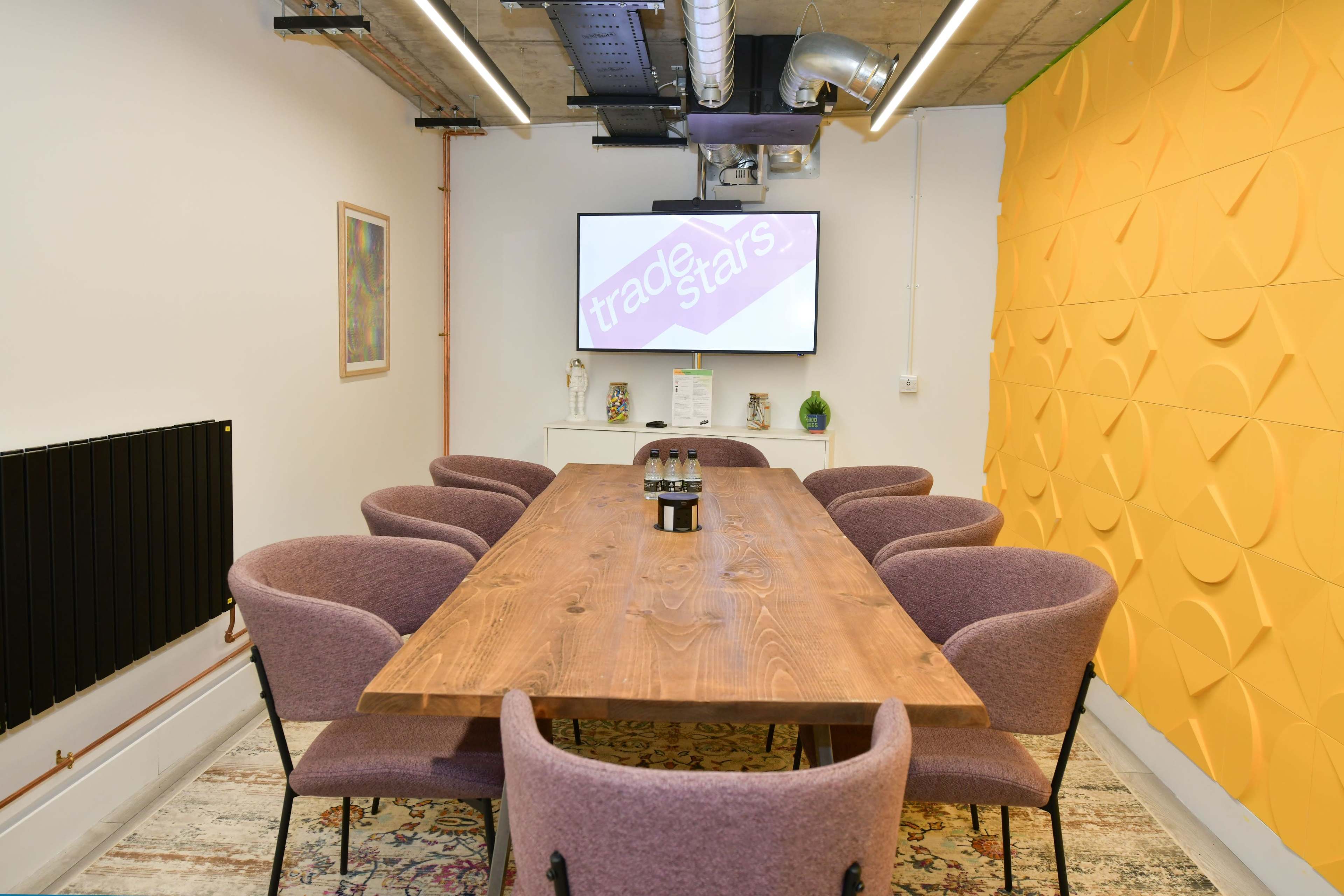 A conference room features a long wooden table surrounded by purple chairs, with a screen mounted on the wall displaying the text "trader stars."