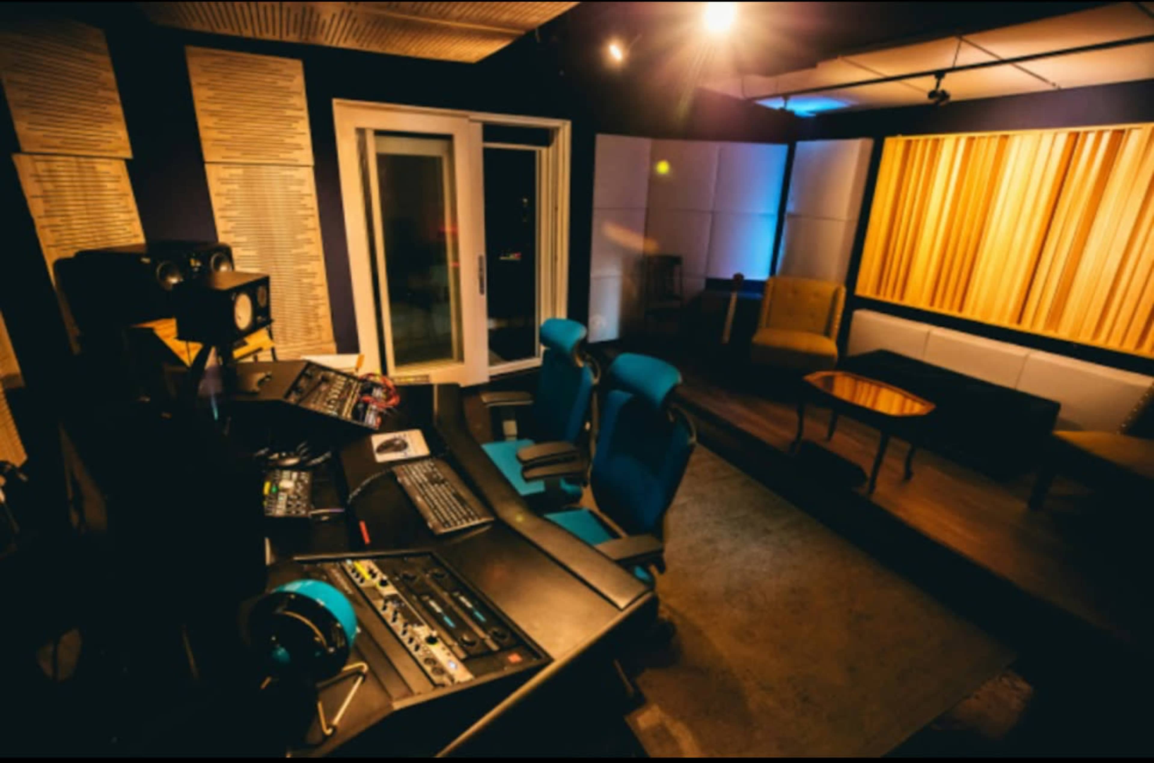 The image shows a recording studio with a control desk, two blue chairs, and a seating area featuring a sofa and a chair.