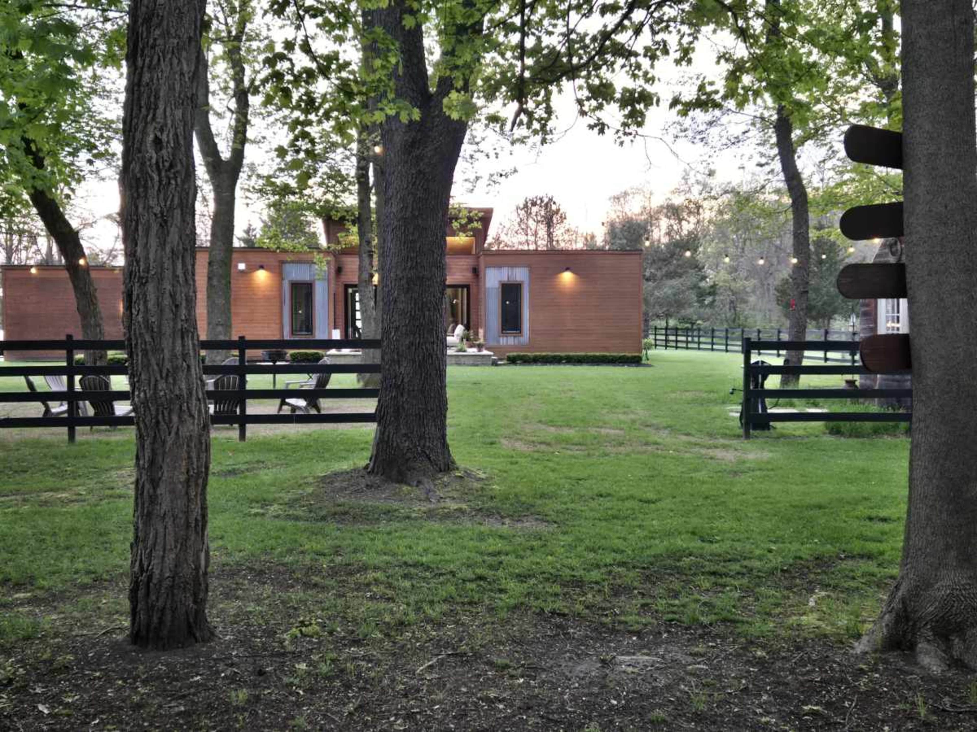 A modern wooden structure is set among trees in a grassy area surrounded by a black fence.