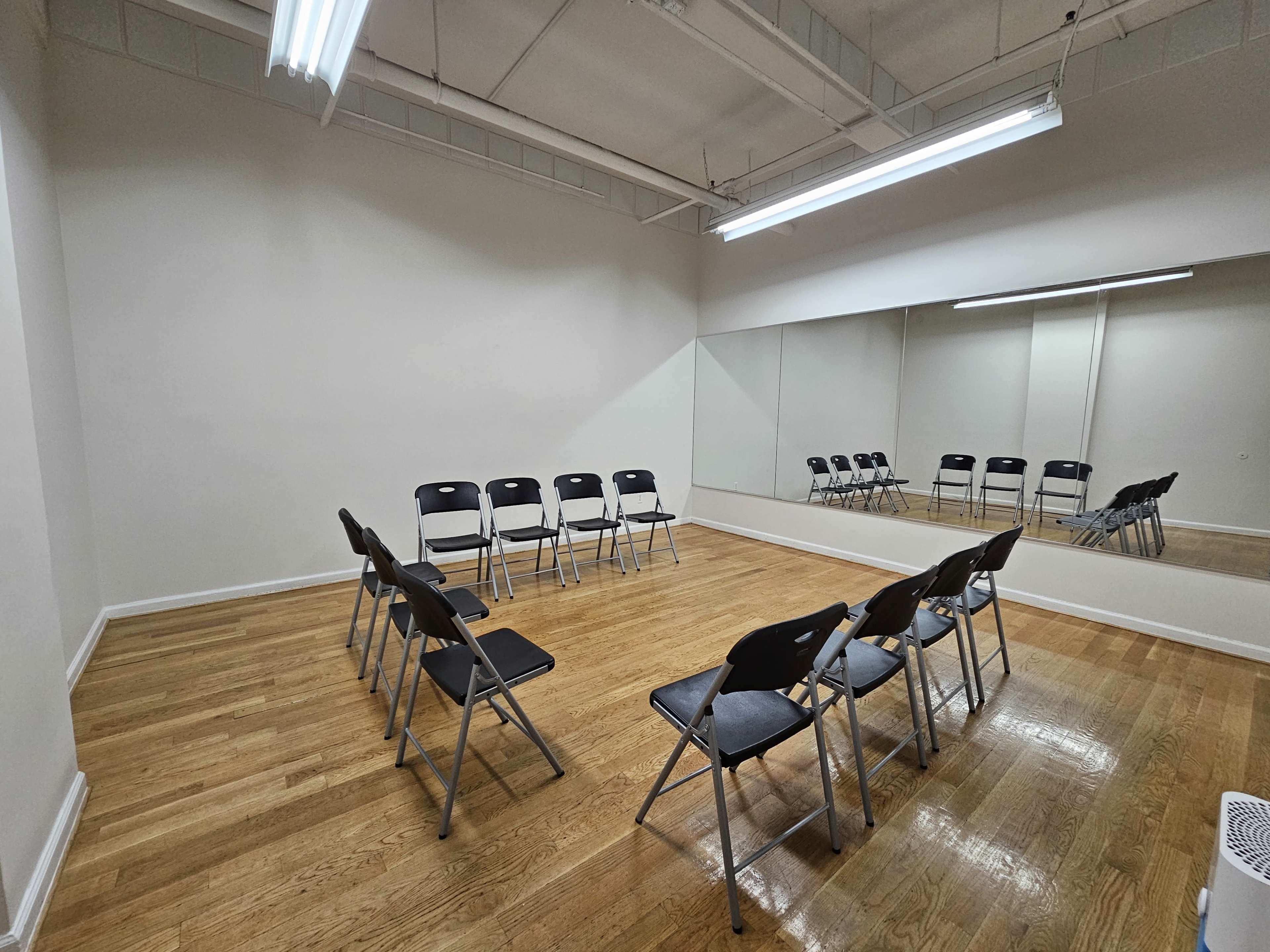 The image shows a spacious, empty room with a row of black folding chairs facing a large mirror on one wall.