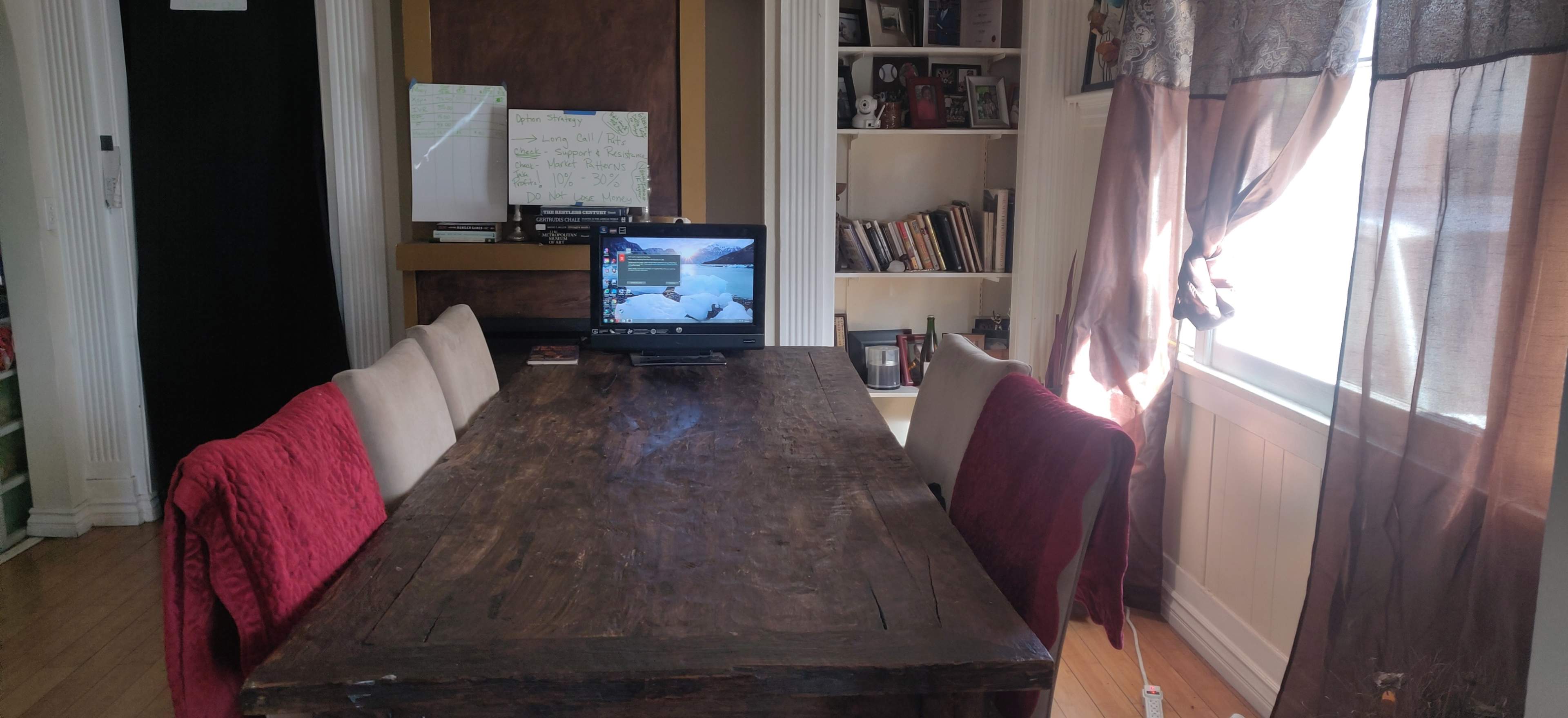 A wooden dining table with six chairs is positioned in a room featuring a television displaying a winter scene, along with shelves filled with books and framed photos.