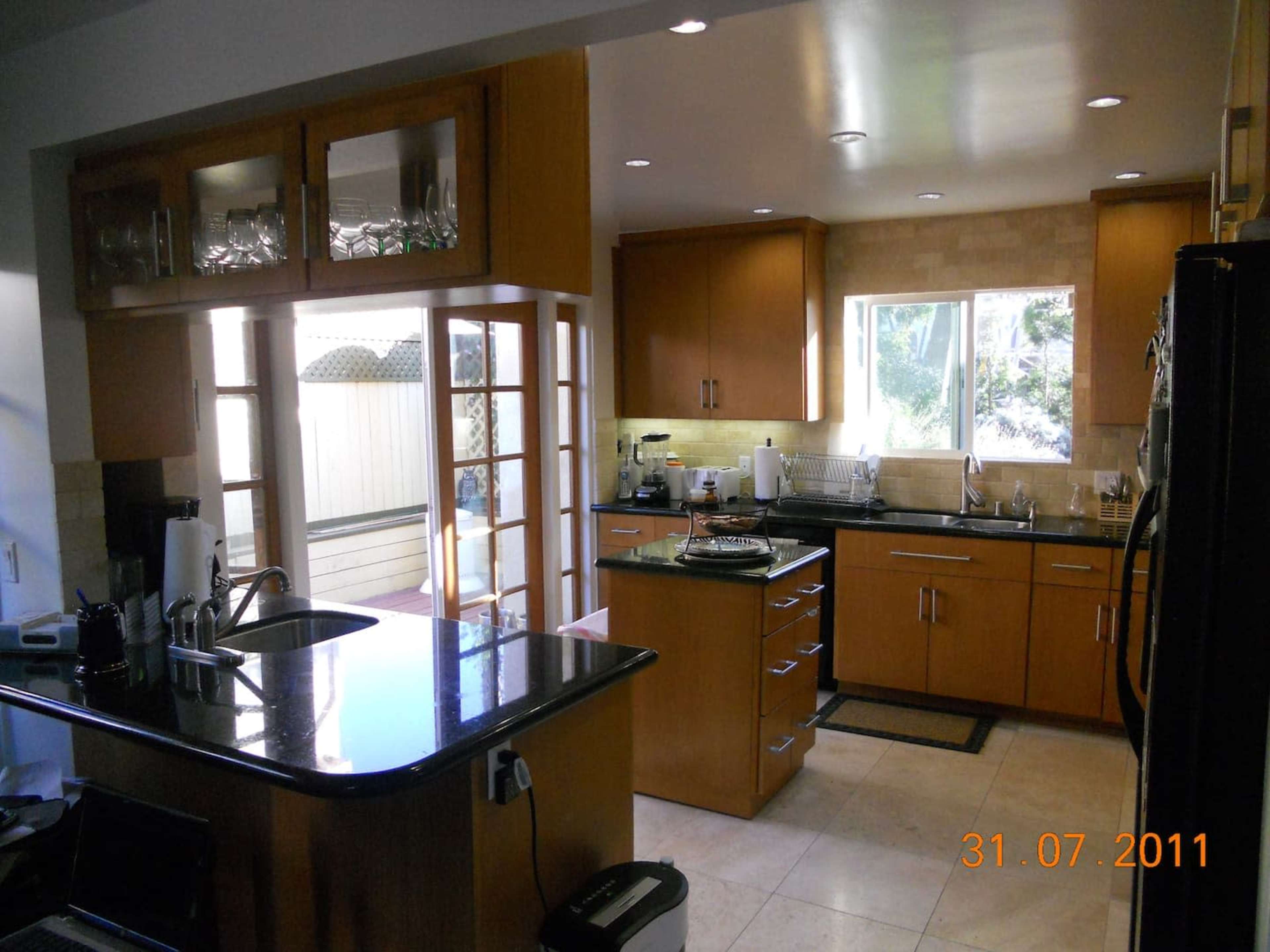 A modern kitchen features wooden cabinets, a central island with a black countertop, and large windows allowing natural light.