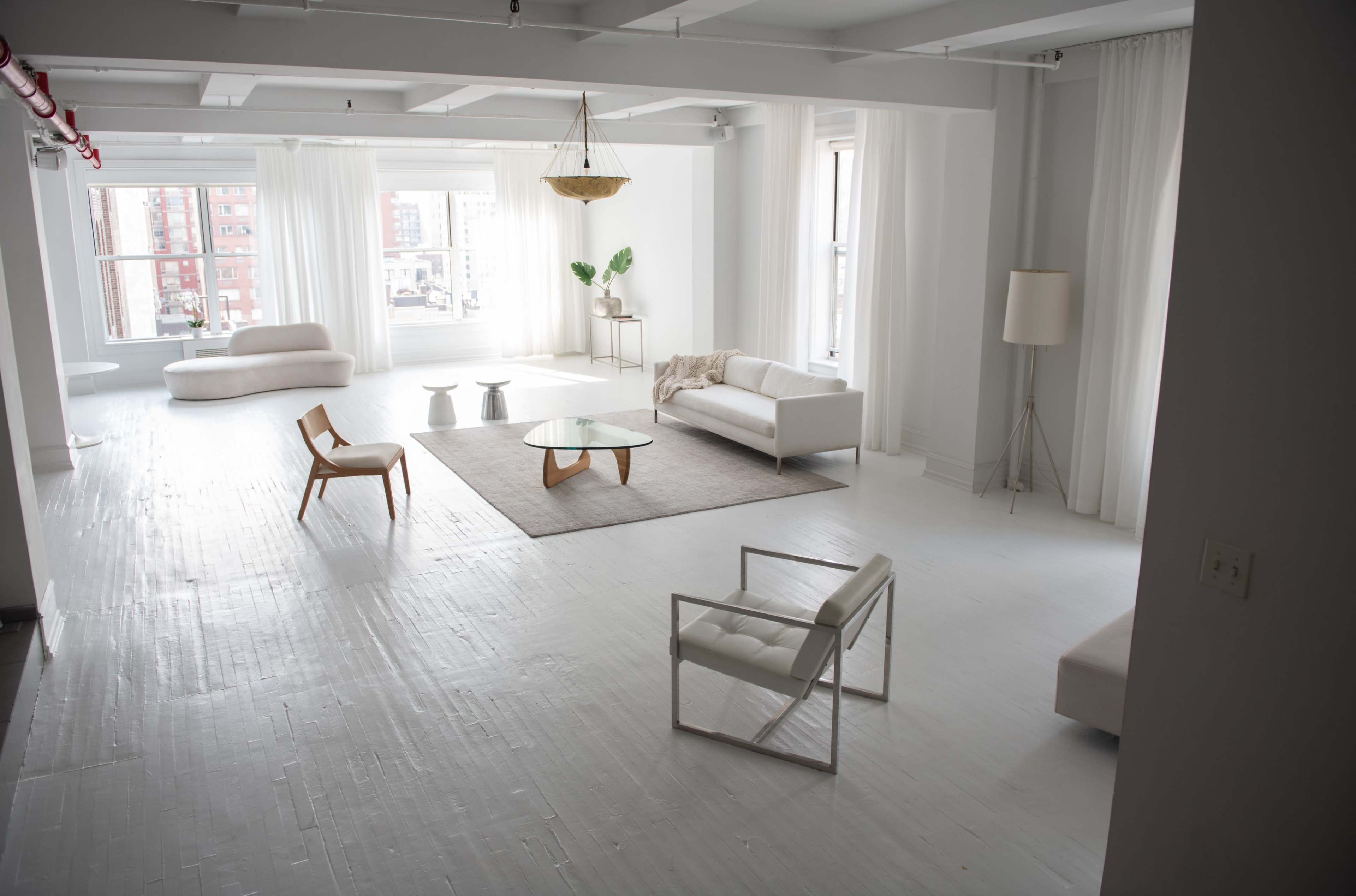 The image shows a spacious, minimalist living room with large windows, white walls, and light-colored furniture arranged around a glass coffee table.