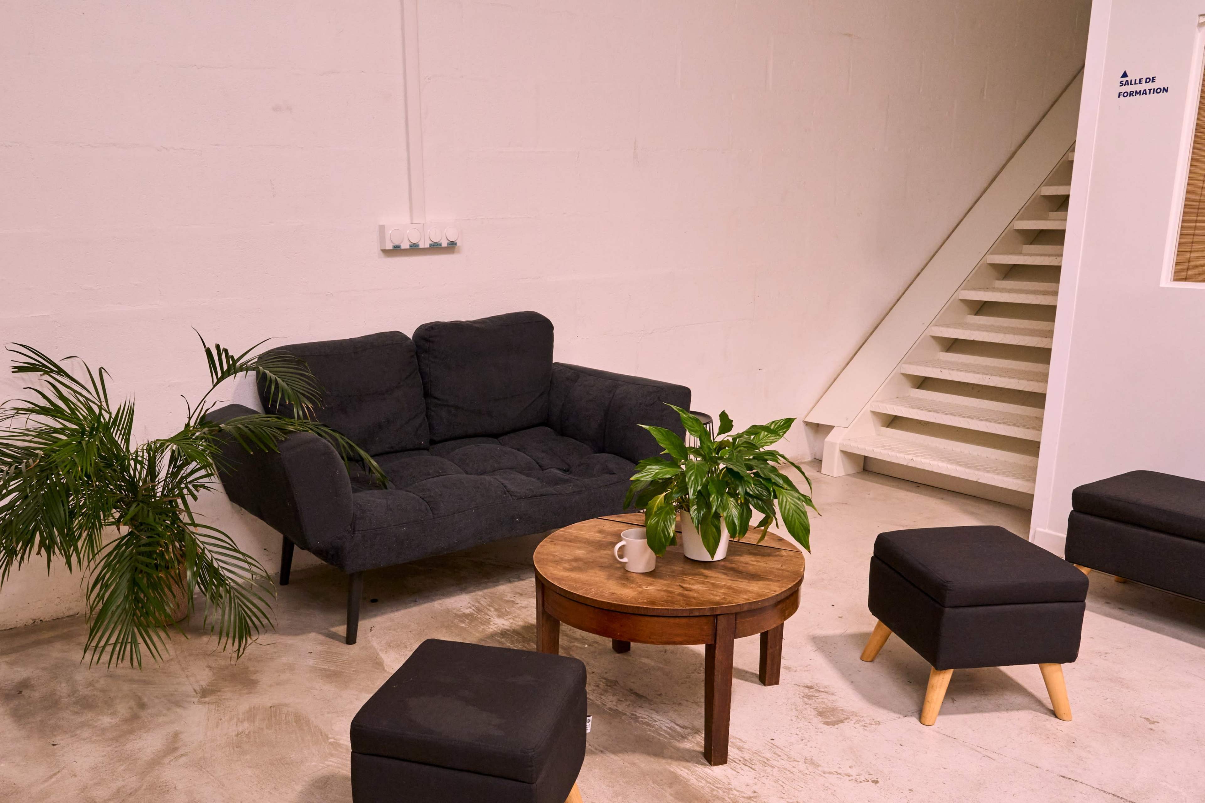 The image shows a minimalist indoor seating area with a dark couch, a round wooden table, potted plants, and a staircase in the background.