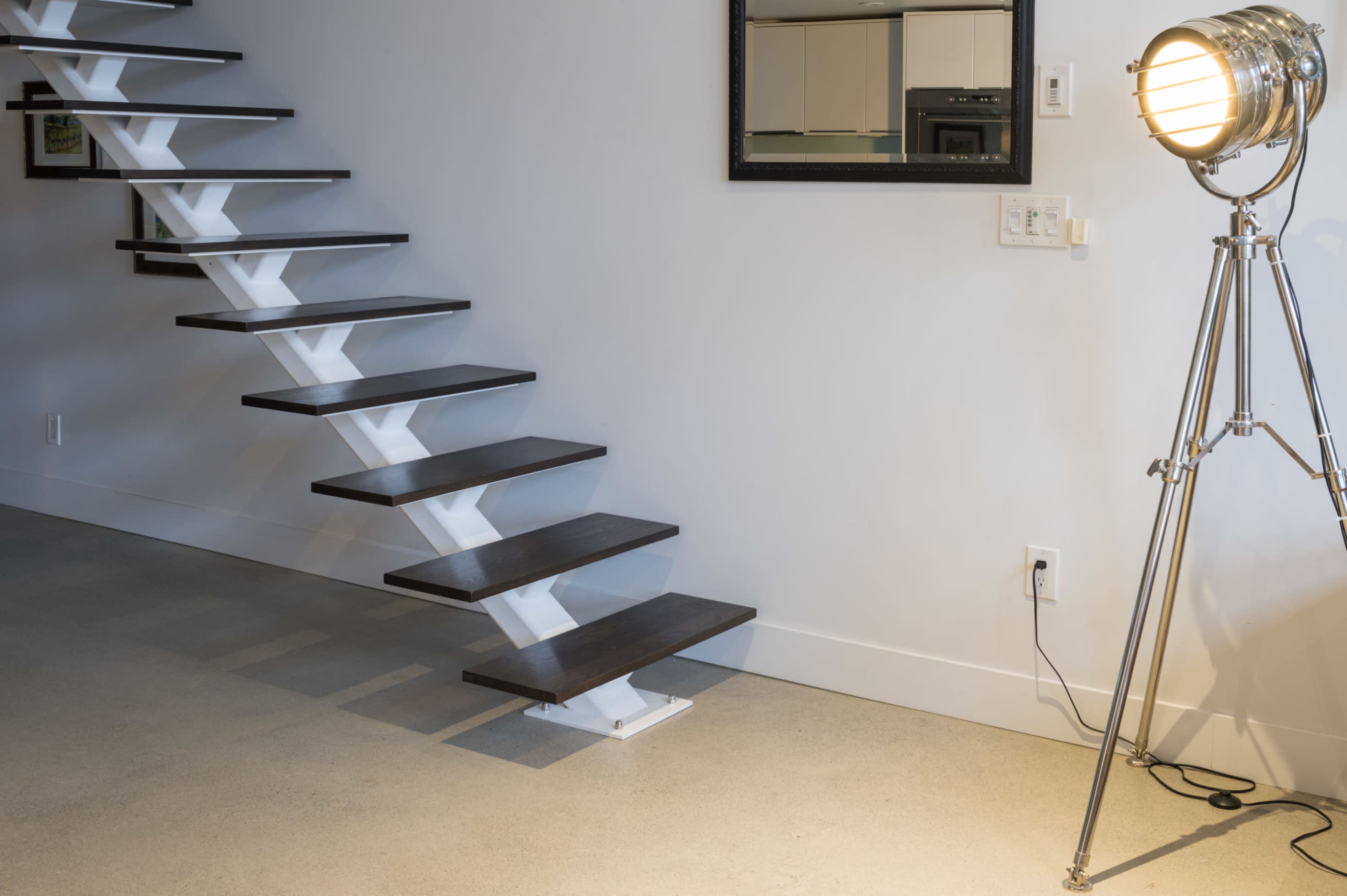 The image shows a modern staircase with wooden steps and a white metal frame, adjacent to a floor lamp on a concrete floor.