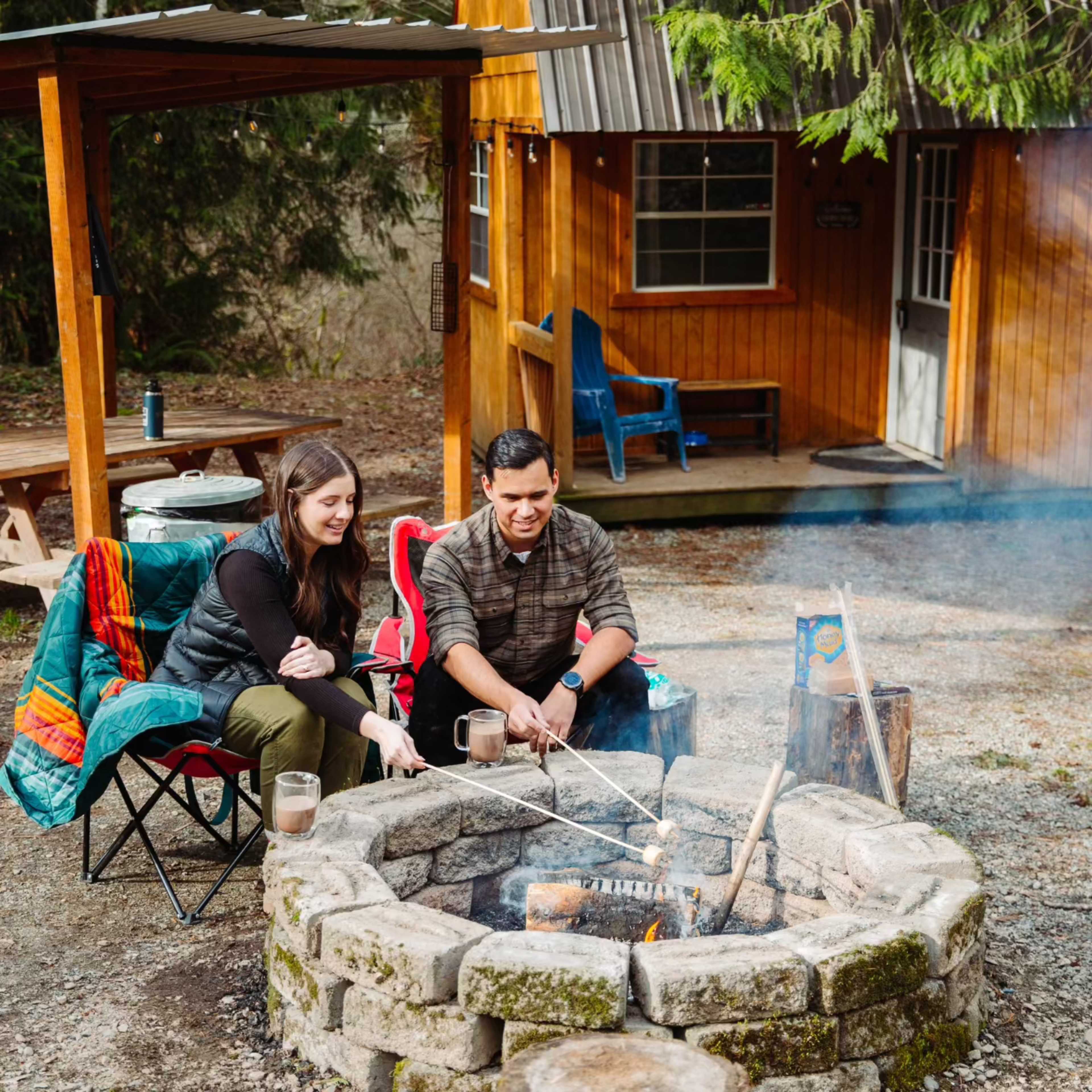 A man and a woman sit by a stone fire pit in front of a cabin, roasting food over the flames.