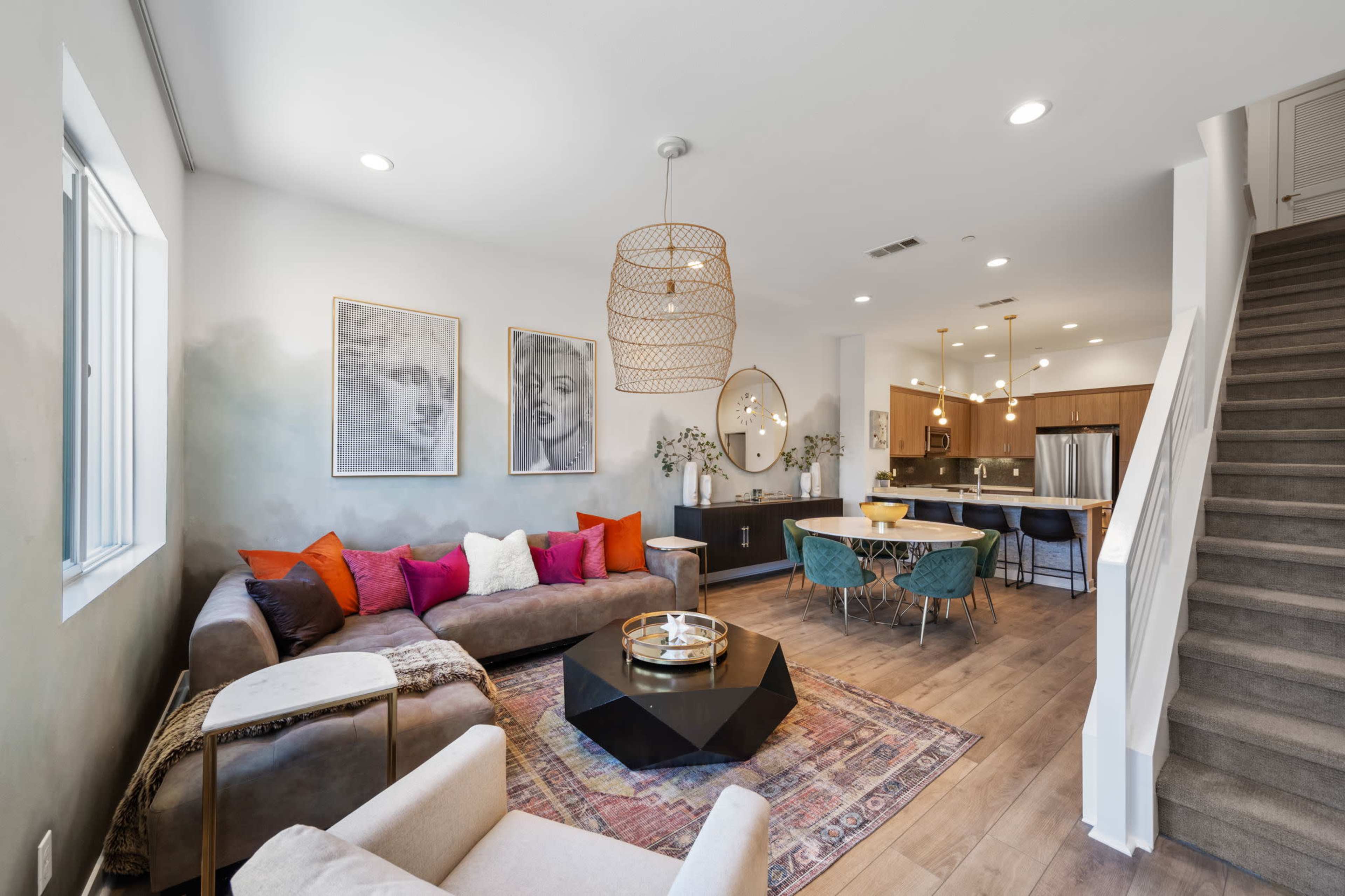 A modern living room features a sectional sofa with colorful cushions, a coffee table, and framed portraits on the walls, leading to a kitchen and staircase in the background.