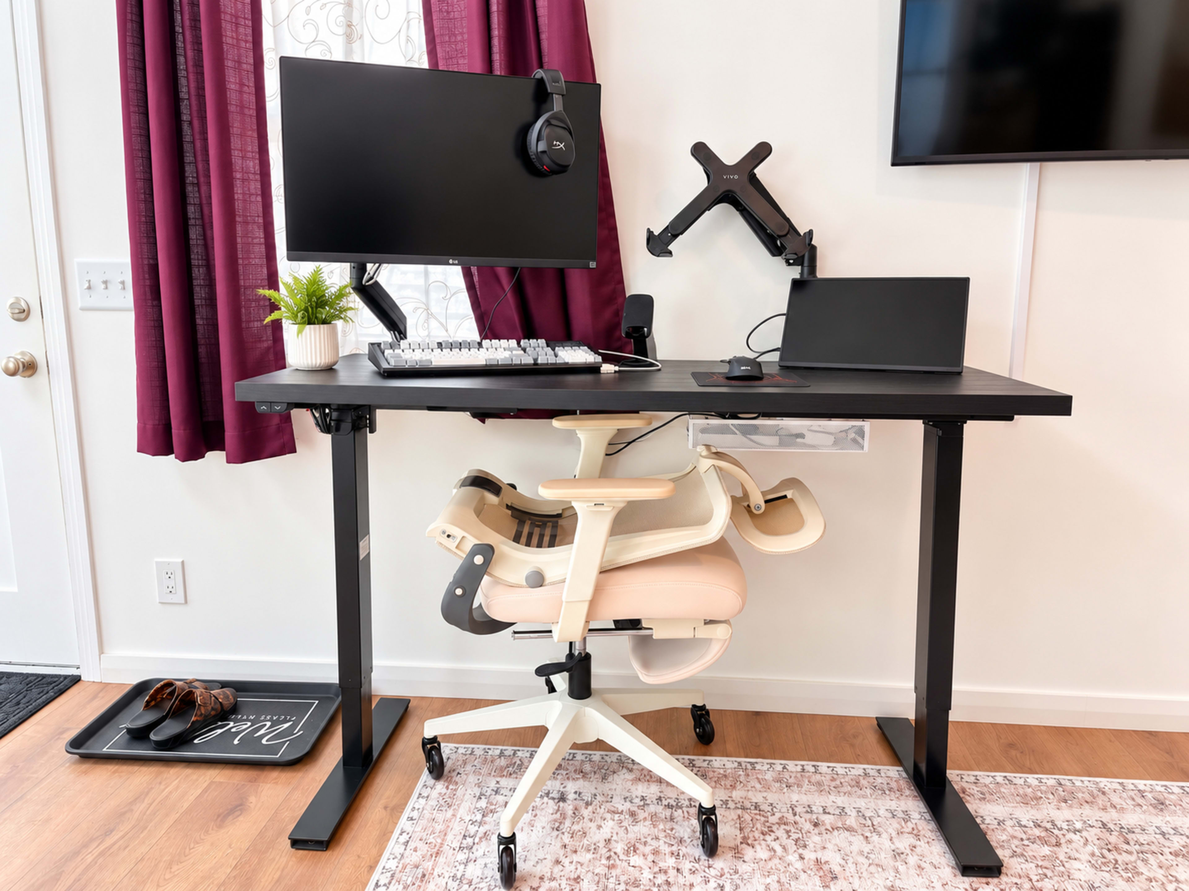 A height-adjustable desk equipped with a monitor, keyboard, headset, and laptop sits beside a chair against a wall with burgundy curtains.