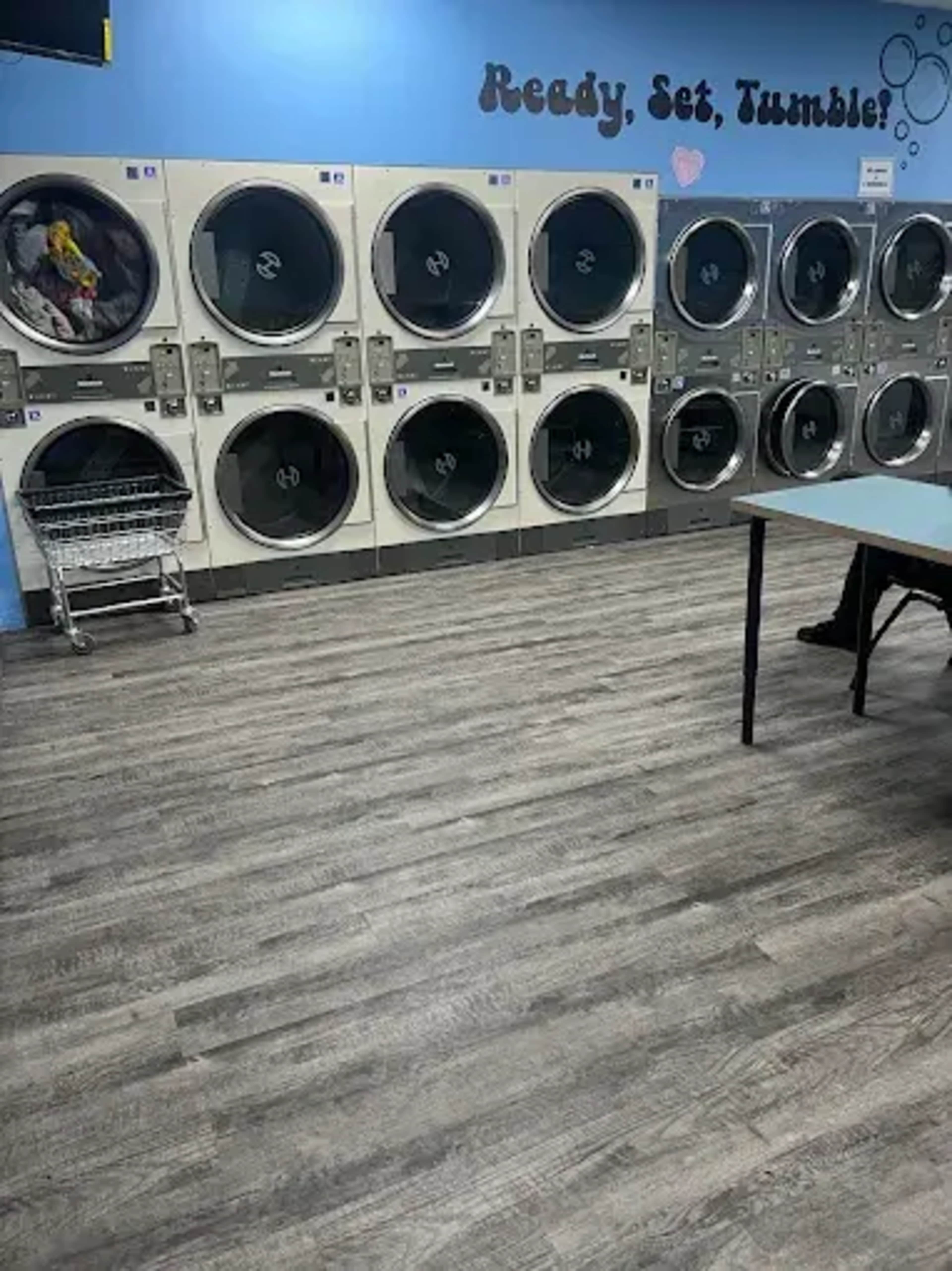 The image shows a laundromat with rows of coin-operated dryers and a table, all situated on a wooden floor.