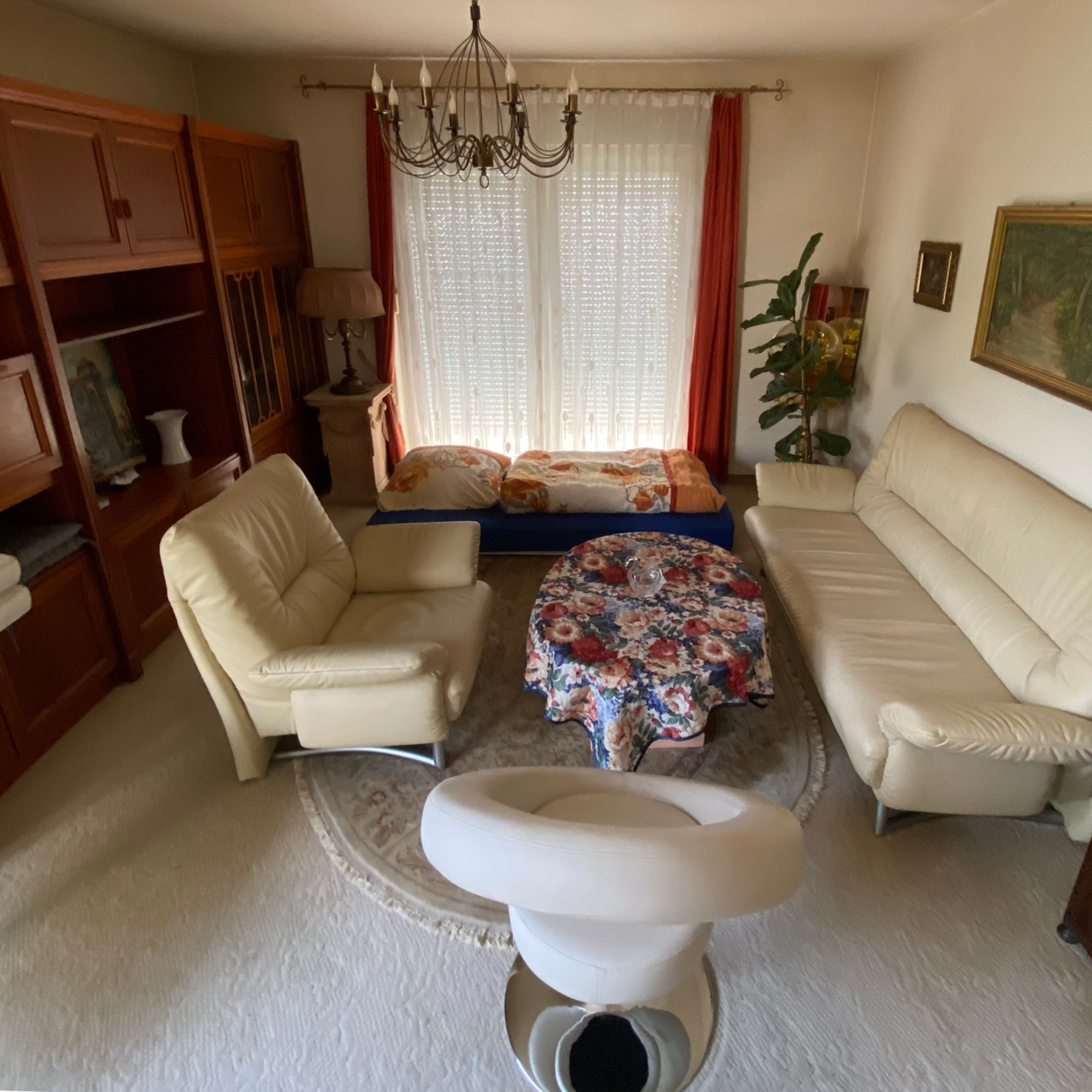 The image shows a living room with two sofas, a round table covered with a floral tablecloth, and a bed in the background near a window.