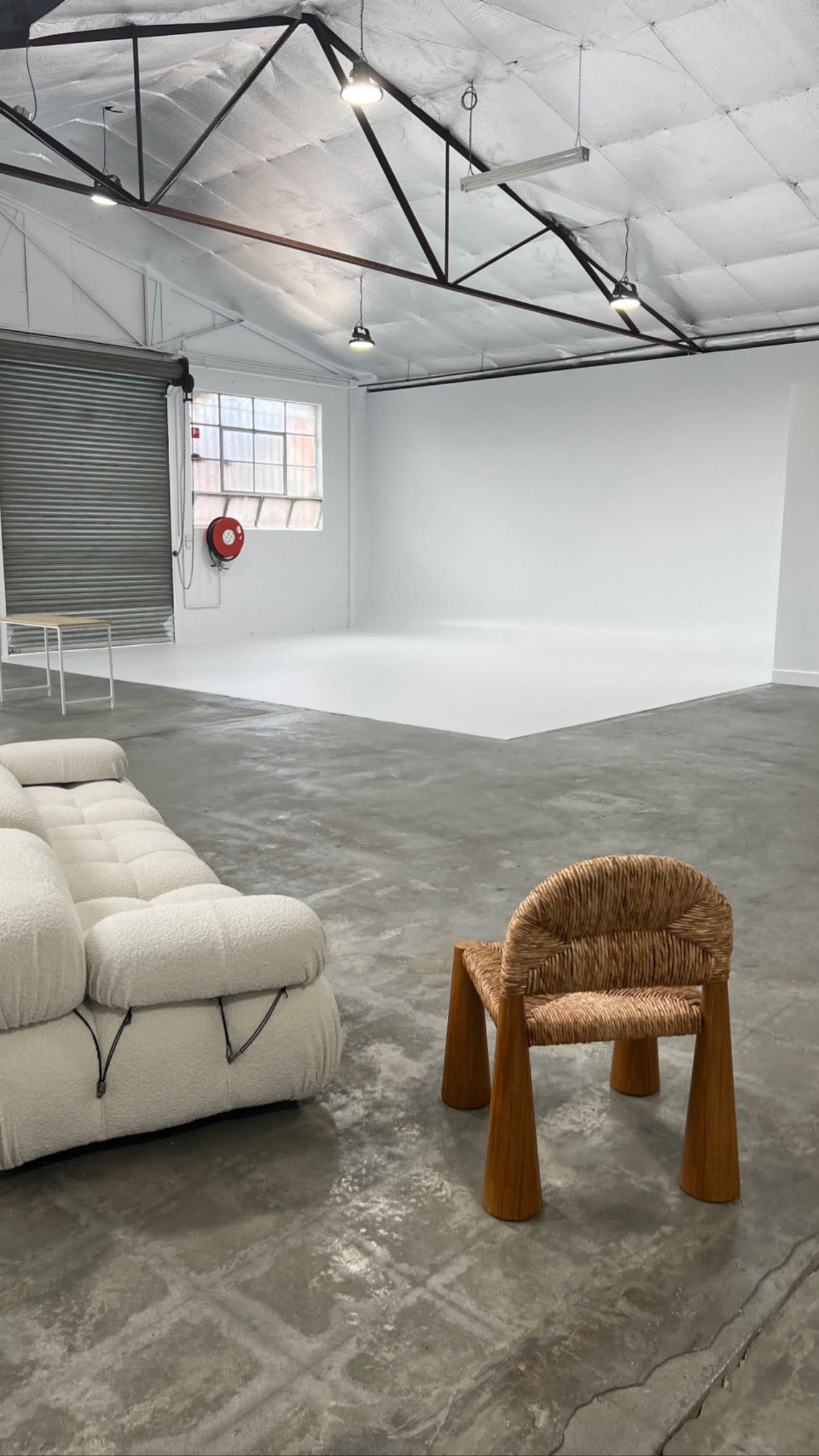 An empty industrial space with a white wall, concrete floor, a white table in the background, a fire extinguisher on the wall, a textured white sofa, and a wooden chair with a woven seat.
