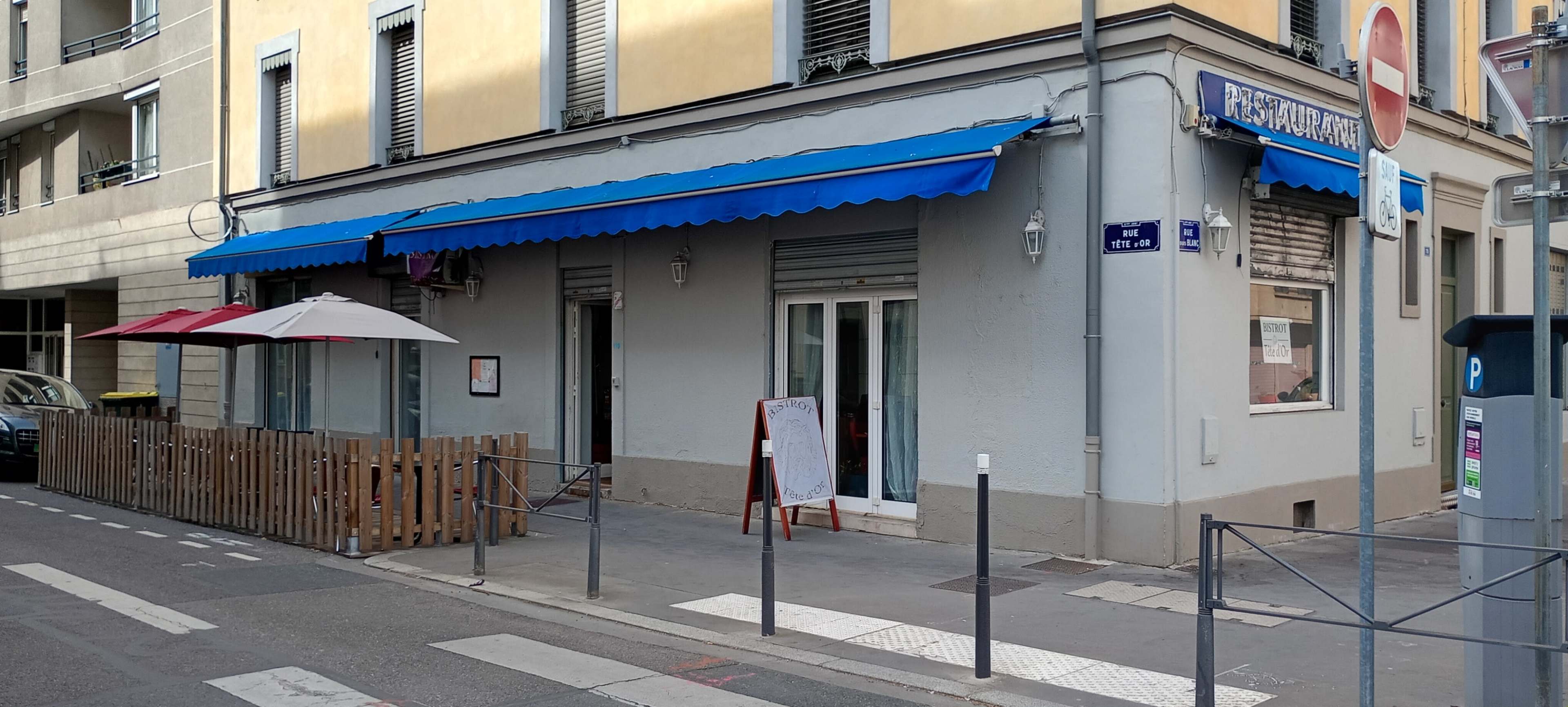 The image shows a restaurant with a blue awning, a fenced outdoor seating area, and a chalkboard menu outside, located on a city street.