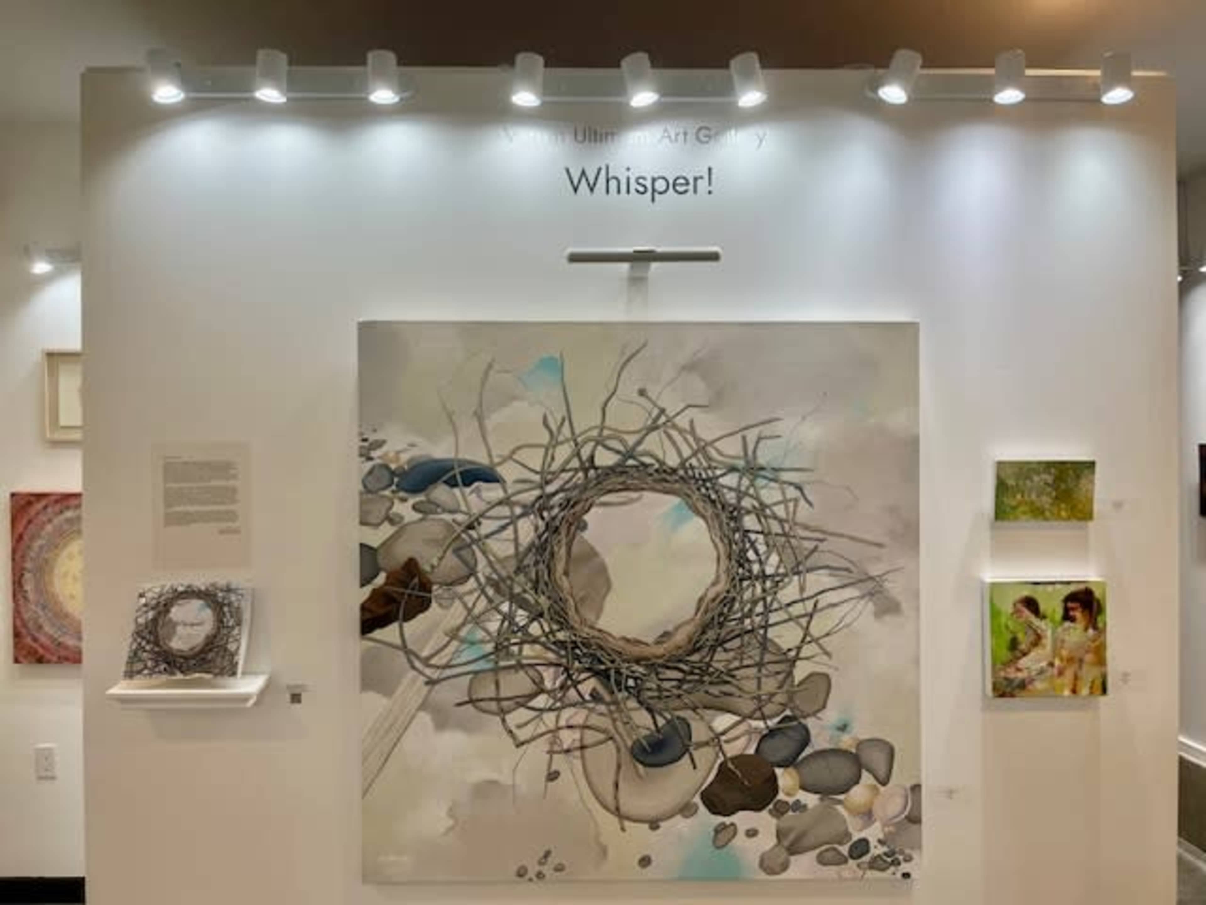 The image shows an art exhibit wall featuring a large painting of a circle made of twigs and stones, with smaller artworks displayed alongside it.