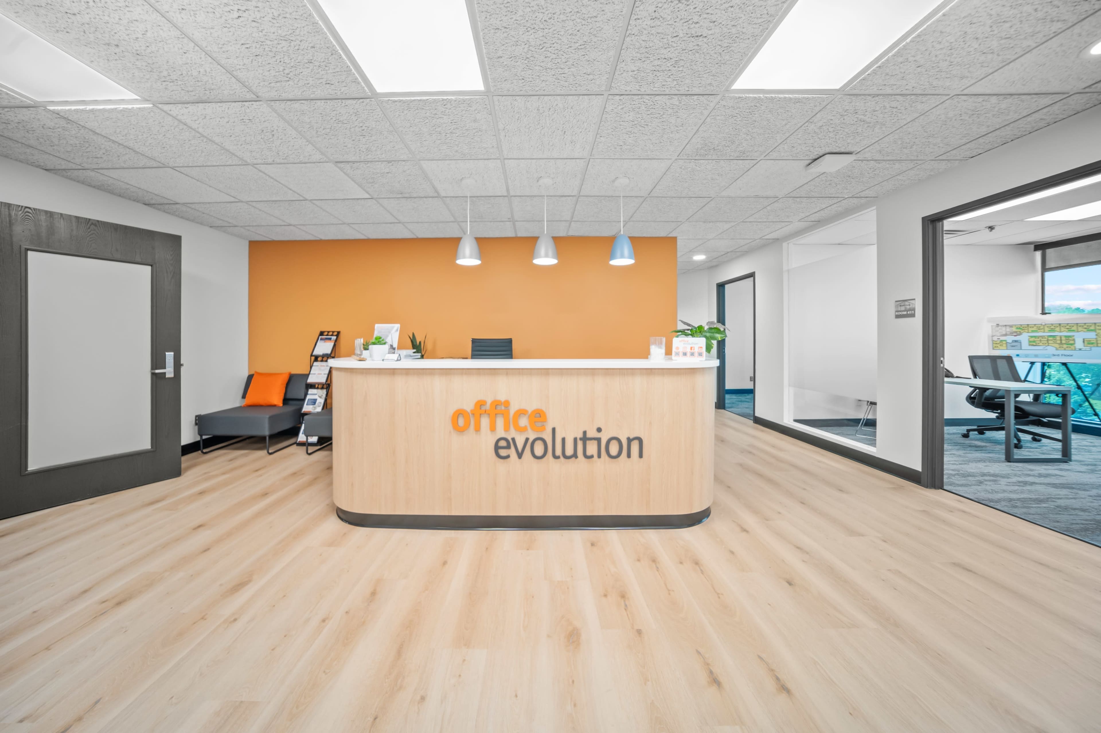 The image shows a modern office reception area with a wooden desk labeled "office evolution" against an orange accent wall, featuring light fixtures and a clean, minimalist design.