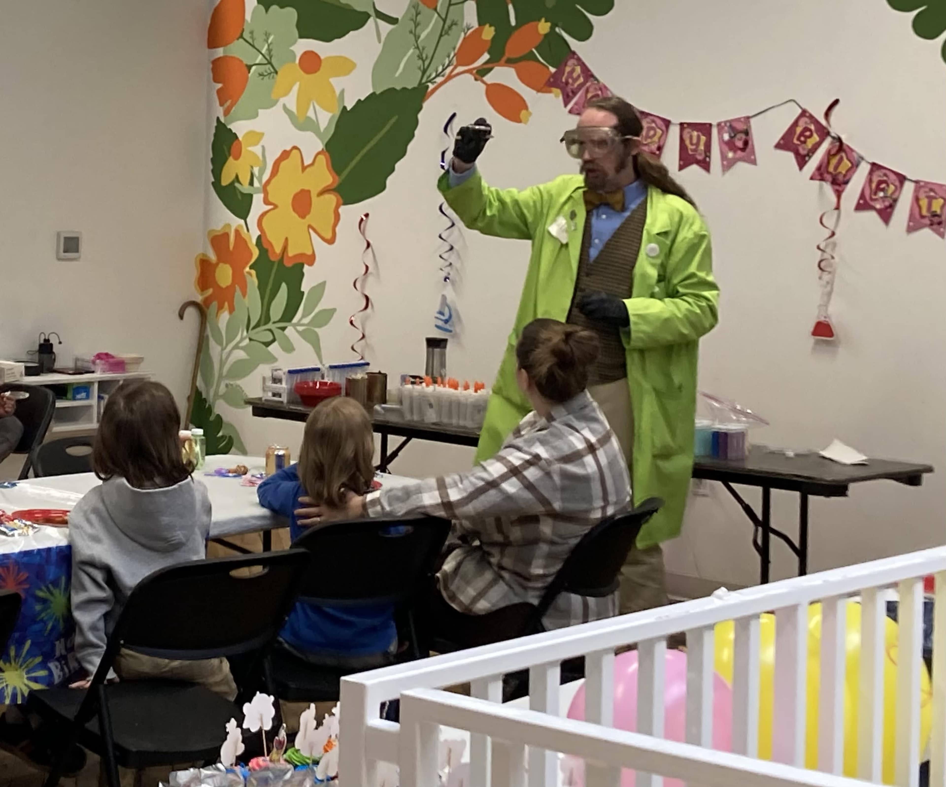 A person in a green lab coat demonstrates an experiment to a group of children seated at a table decorated for a celebration.