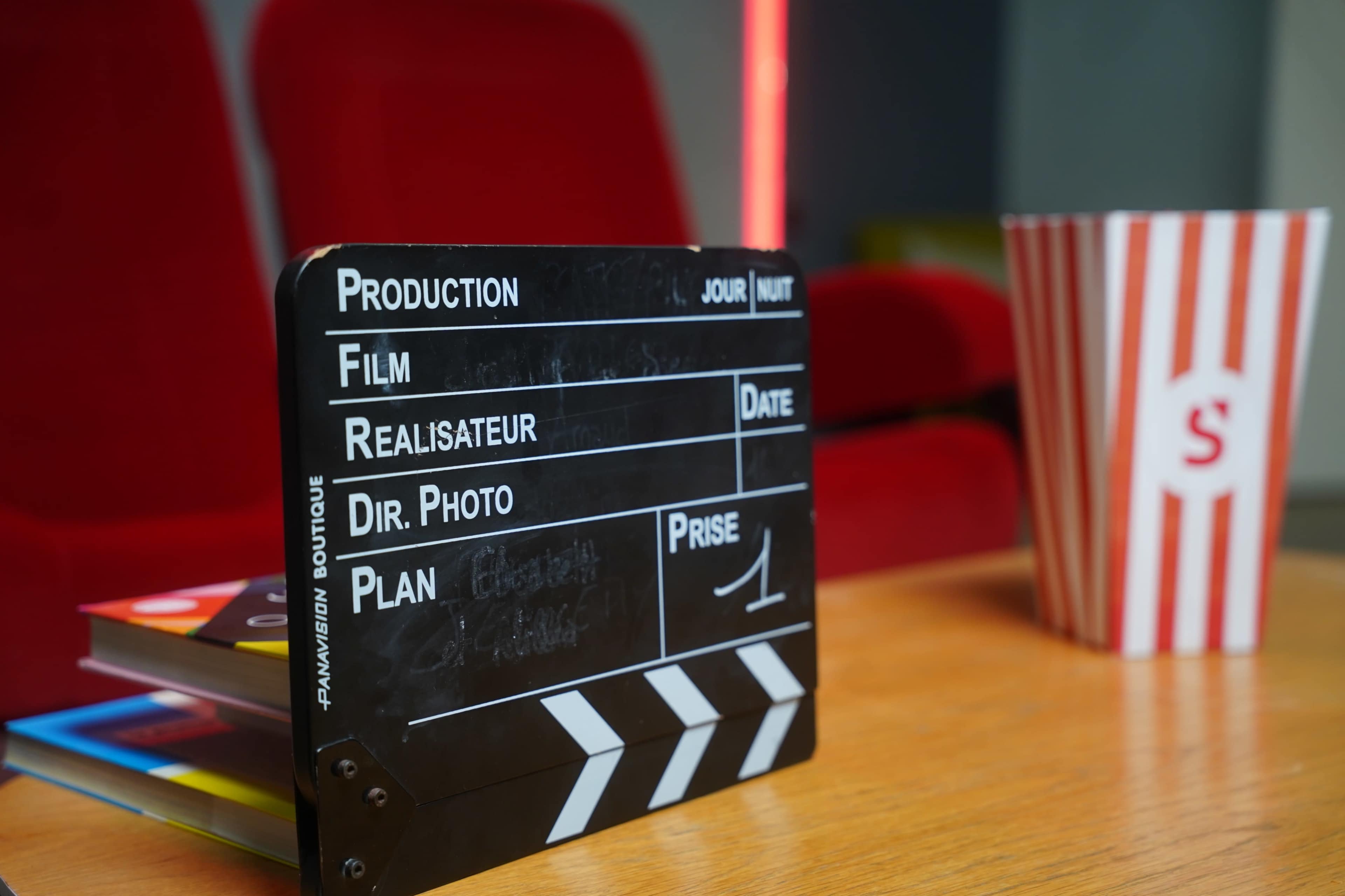 A film clapperboard sits on a wooden table next to a striped popcorn container and colorful books, with red chairs in the background.