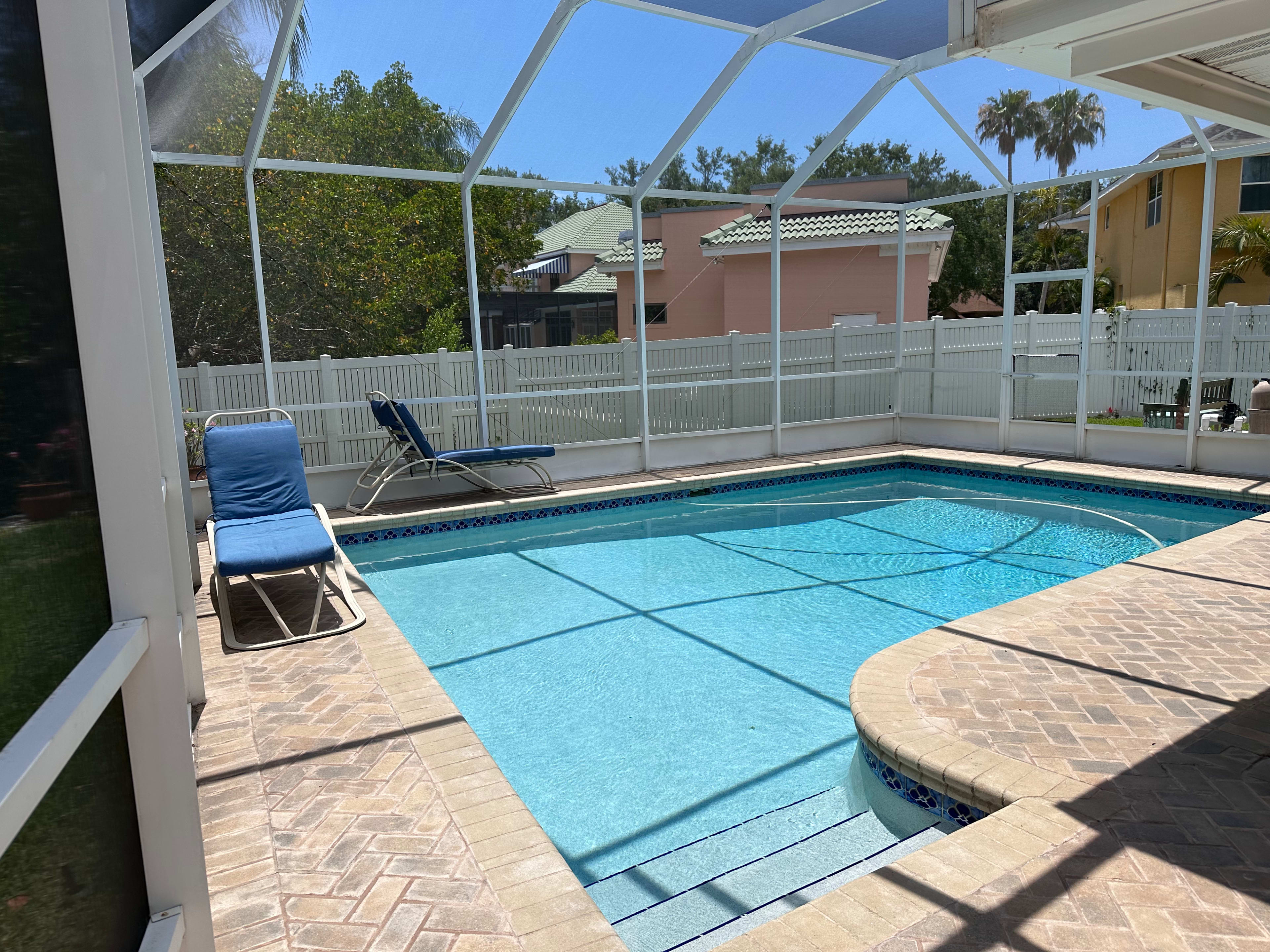 A blue swimming pool is enclosed by a screened patio, featuring a lounge chair and surrounded by greenery and residential buildings.
