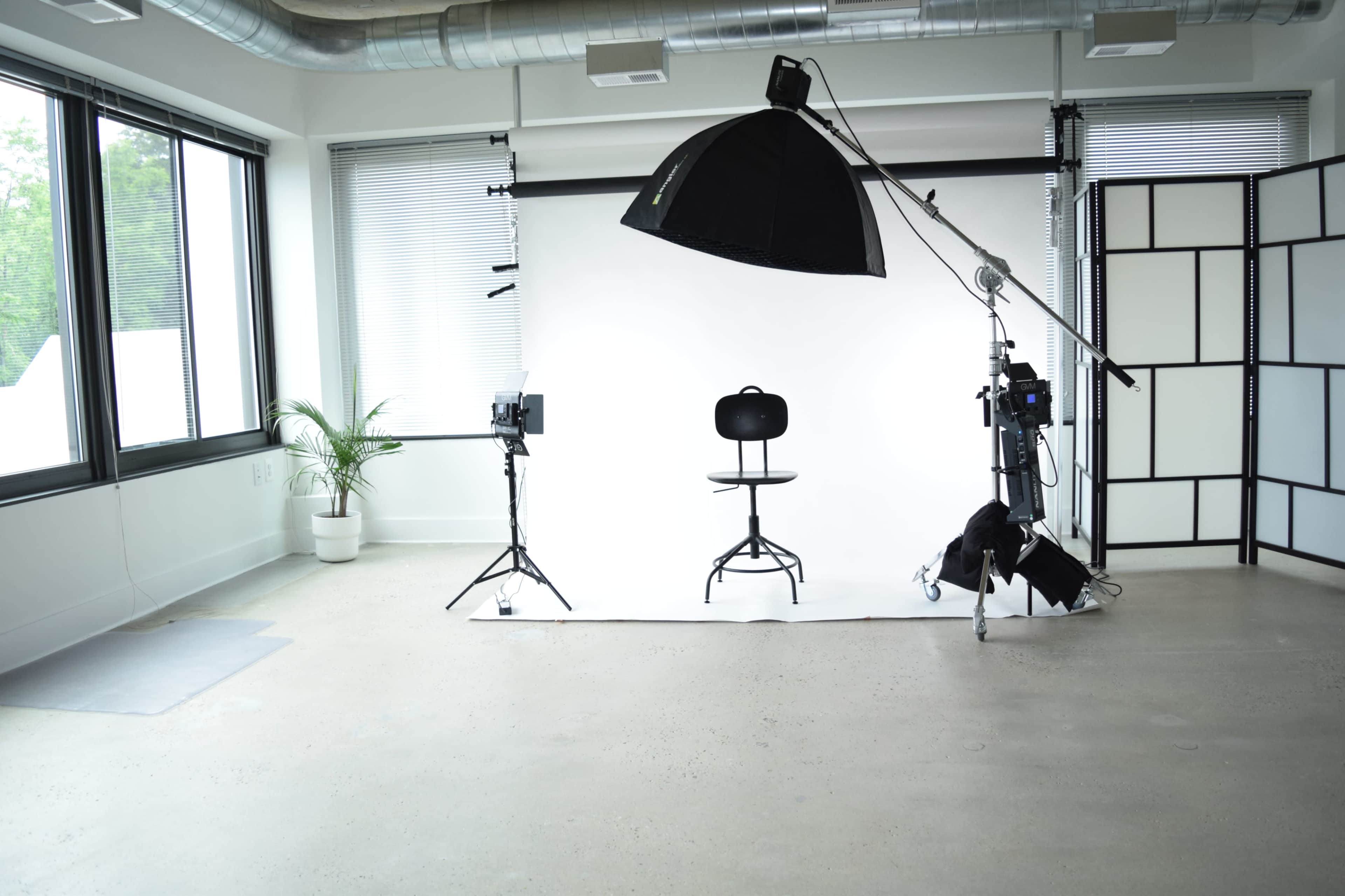 A photography studio featuring a white backdrop, a black stool, studio lighting equipment, and large windows with greenery outside.