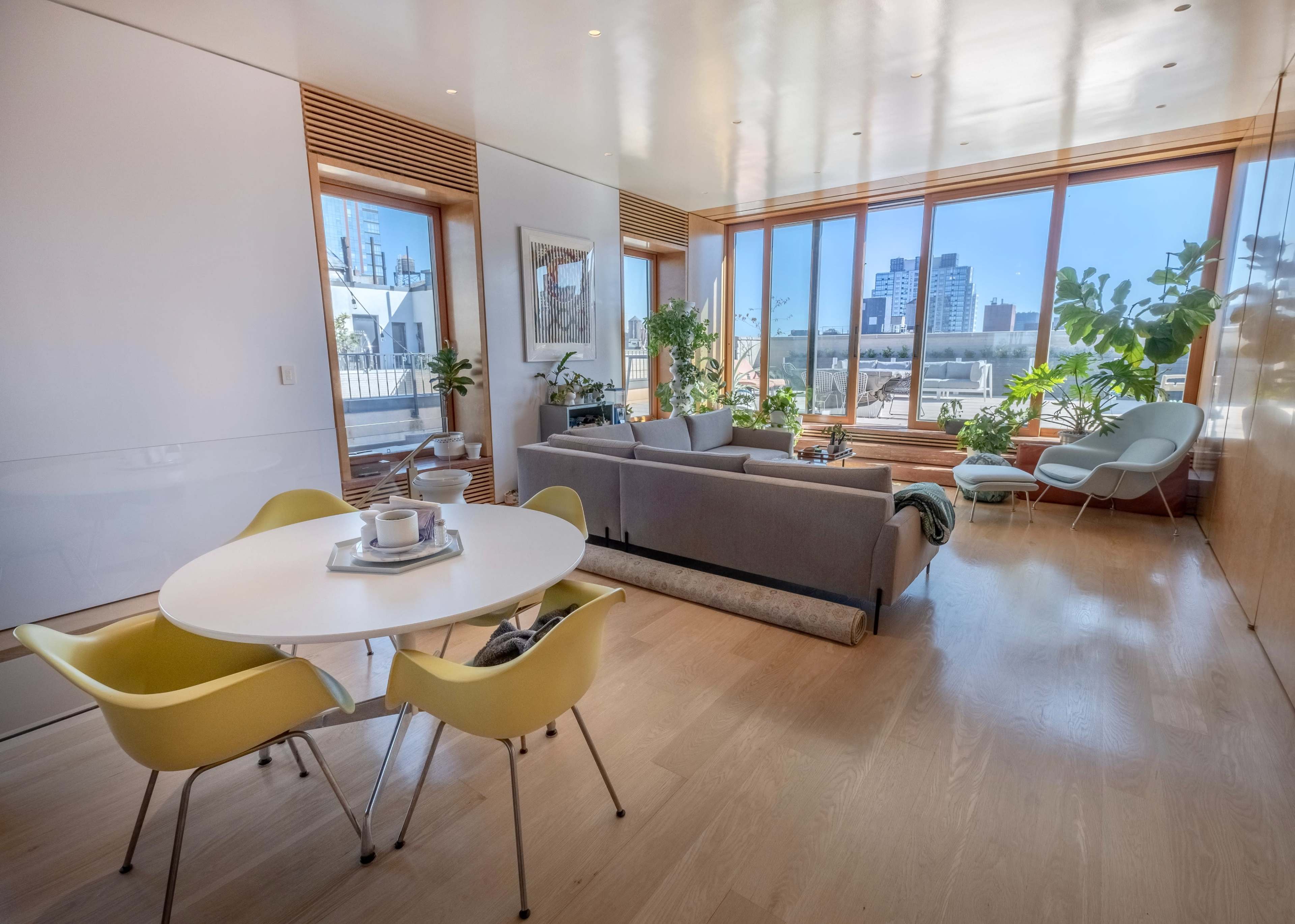 A modern living space features a circular dining table with yellow chairs, a gray sofa, and large windows that offer a view of the city skyline and indoor plants.