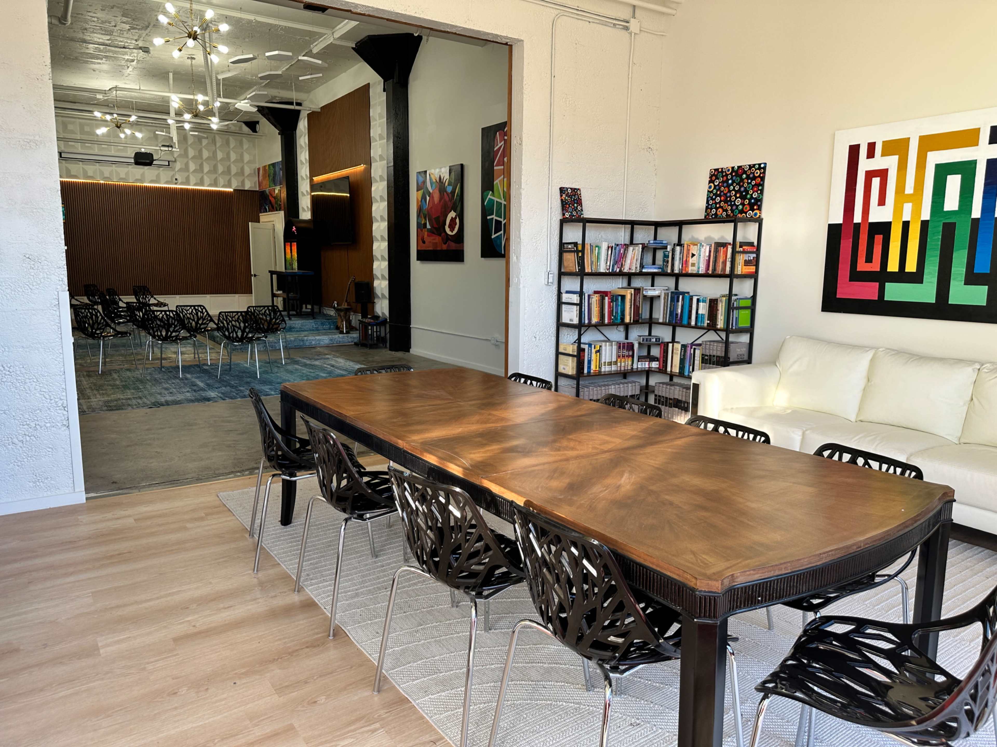 The image shows a modern conference room featuring a long wooden table surrounded by black chairs, with colorful artwork on the walls and a doorway leading to a larger, separate meeting area.
