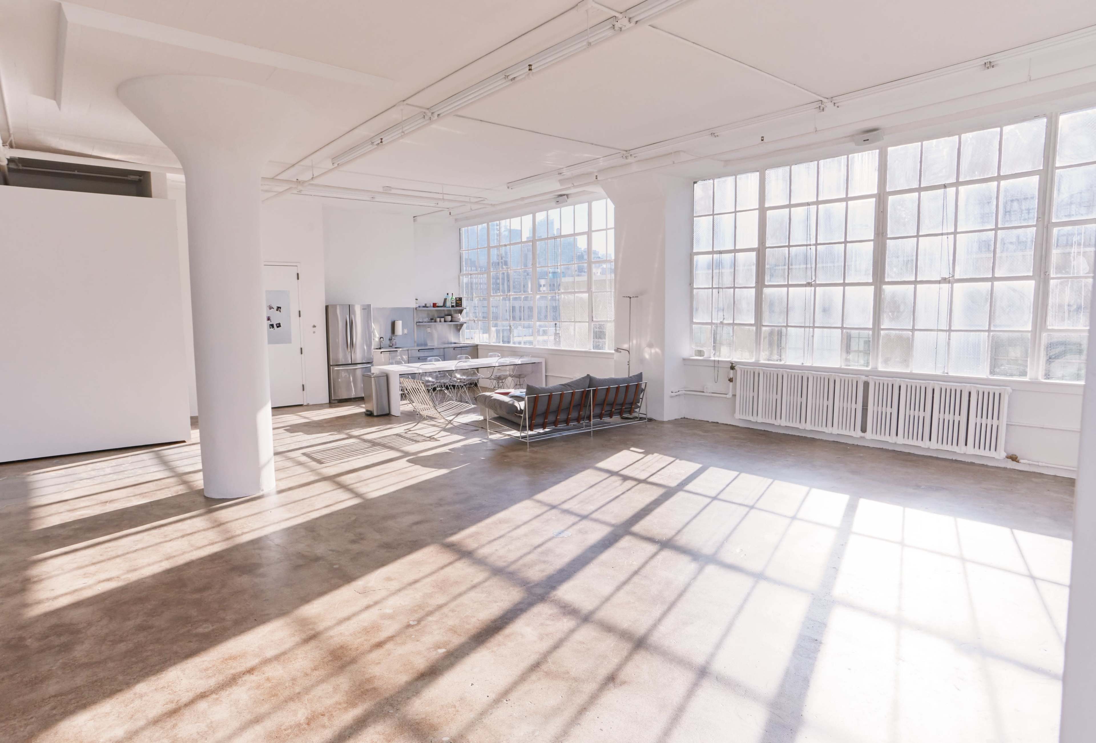 The image shows a spacious, well-lit industrial-style room with large windows, concrete flooring, and minimalist furnishings.