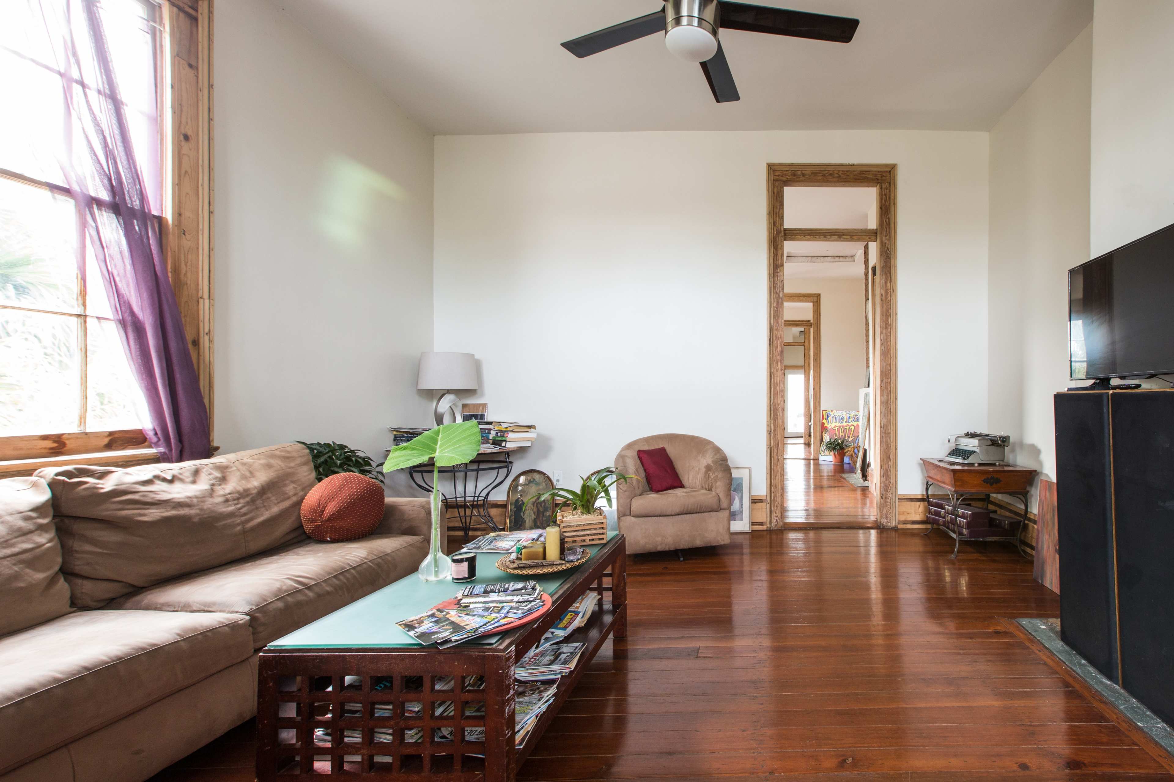 A living room with a beige sofa, a glass-topped coffee table stacked with magazines, and a large mirror reflecting a hallway.