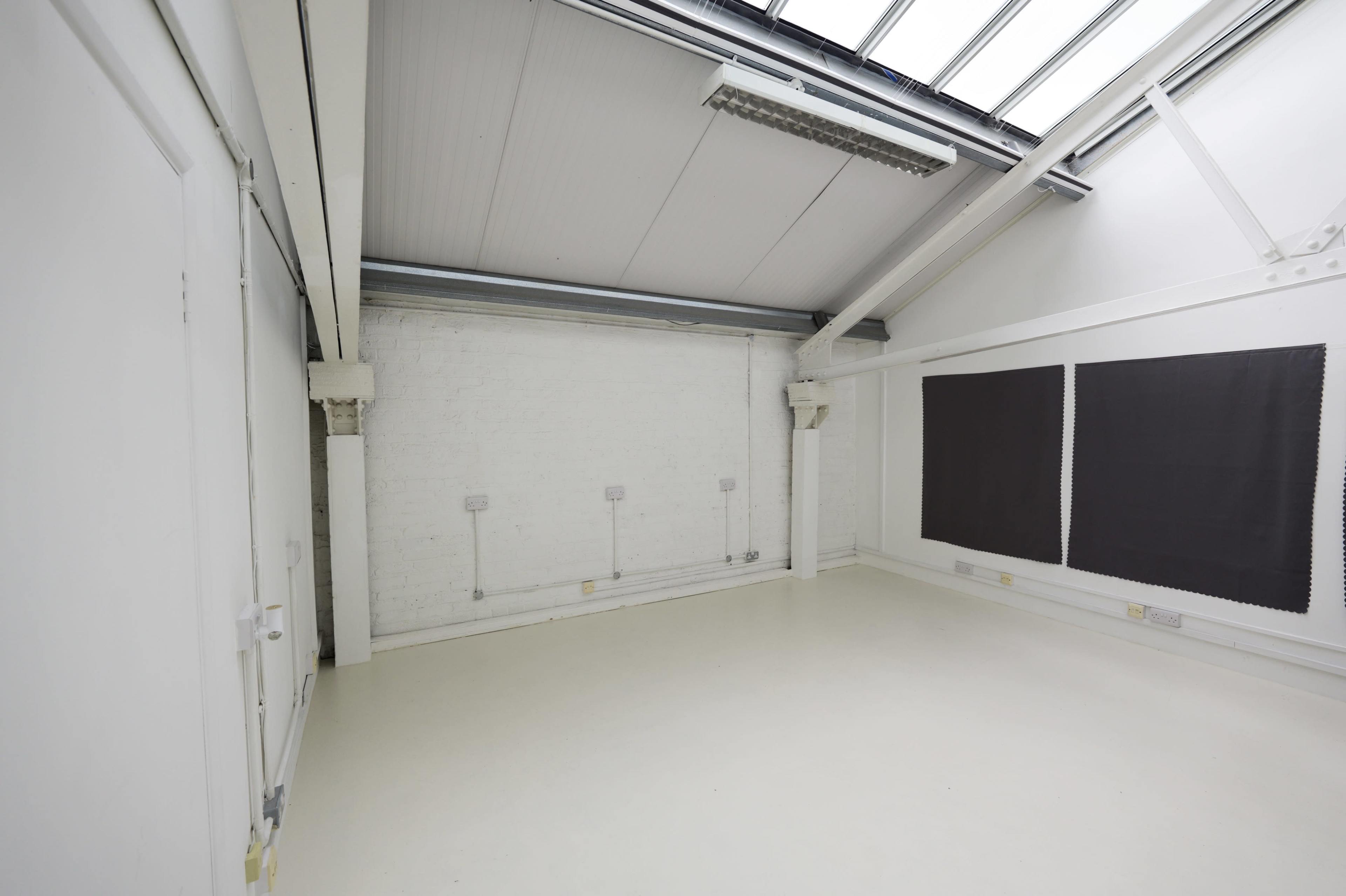 The image shows a large, empty room with white walls, a high ceiling, and two dark panels on one side.