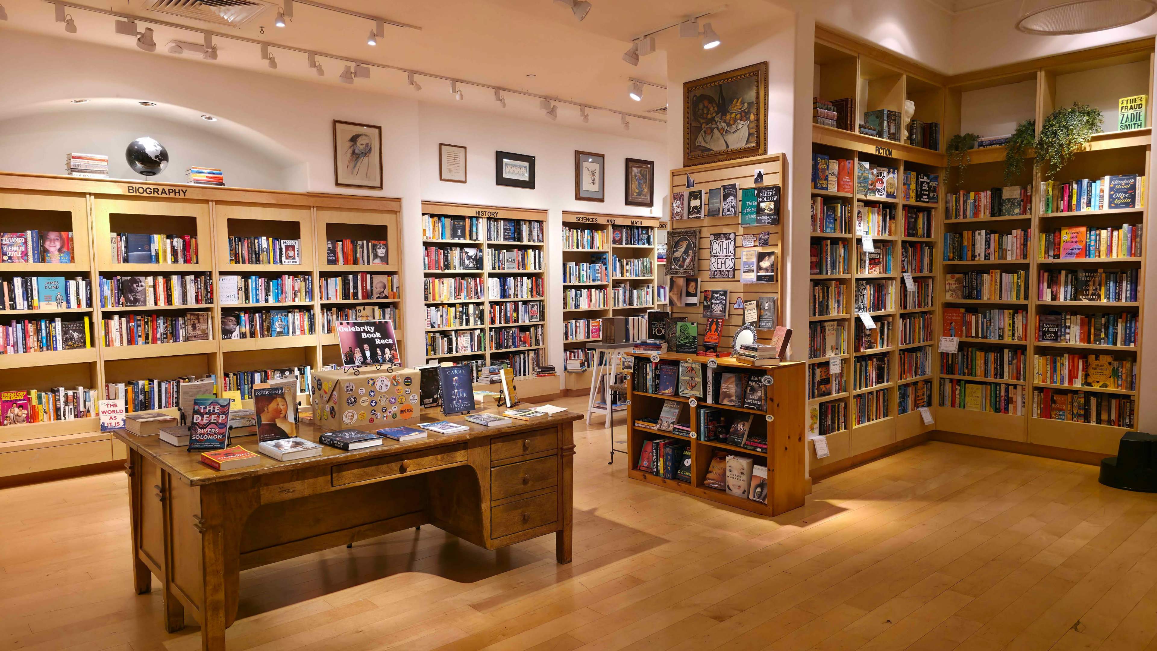 The image shows a cozy bookstore interior with wooden shelves filled with books and a central table displaying various book-related items.