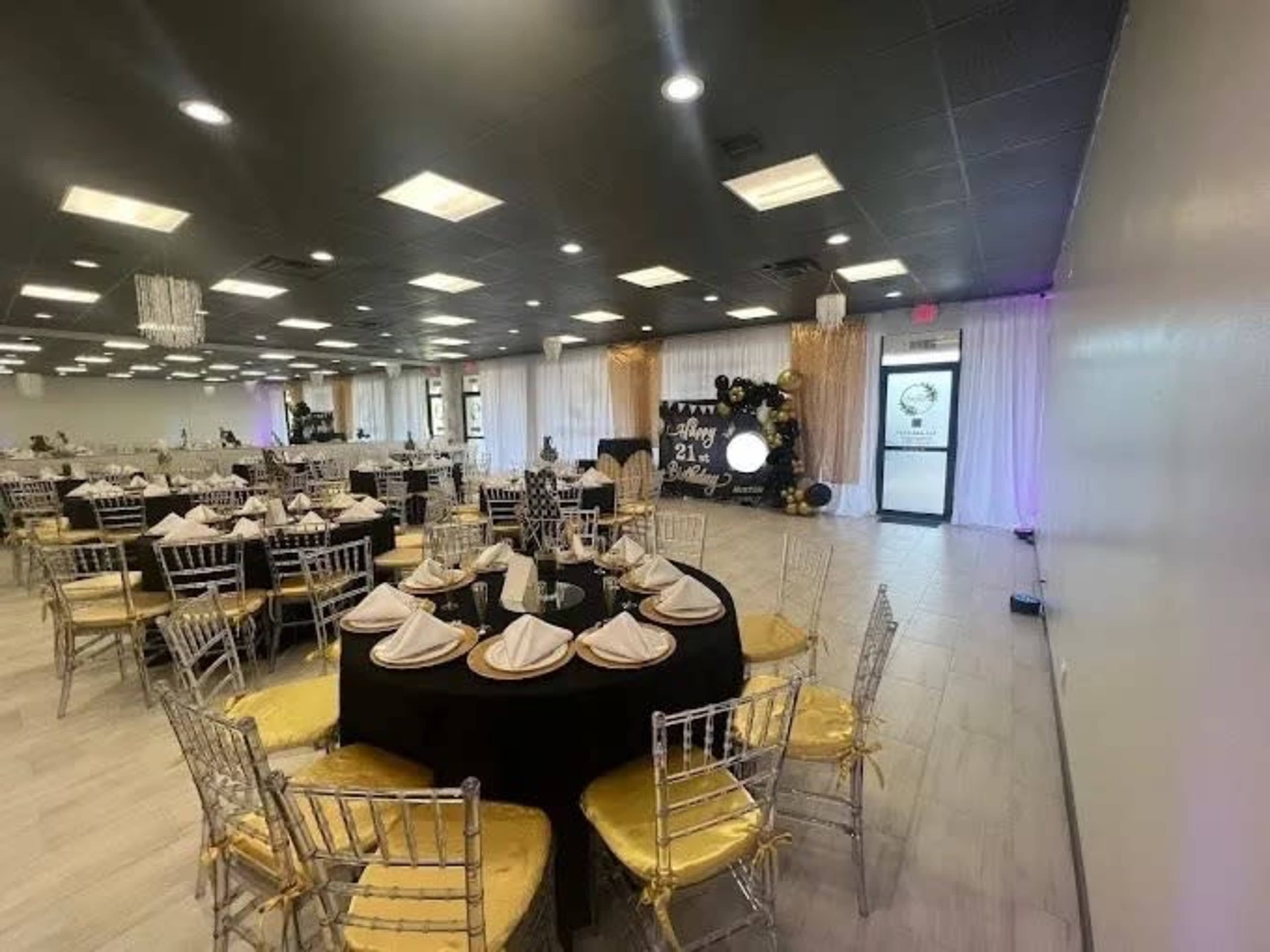 The image shows a banquet hall set up for an event, featuring round tables with black and gold tablecloths, elegant chairs, and decorative elements including curtains and wall decor.