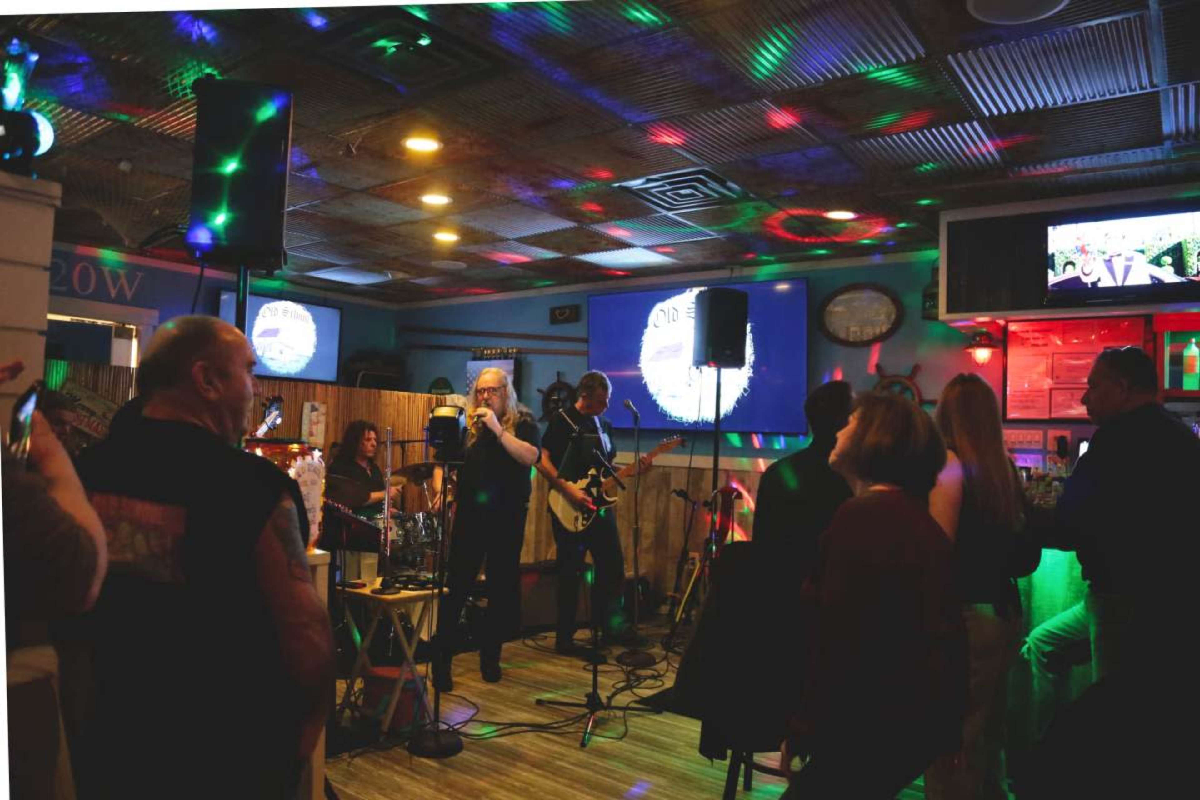 A band performs on stage in a lively bar while colorful lights illuminate the crowd.