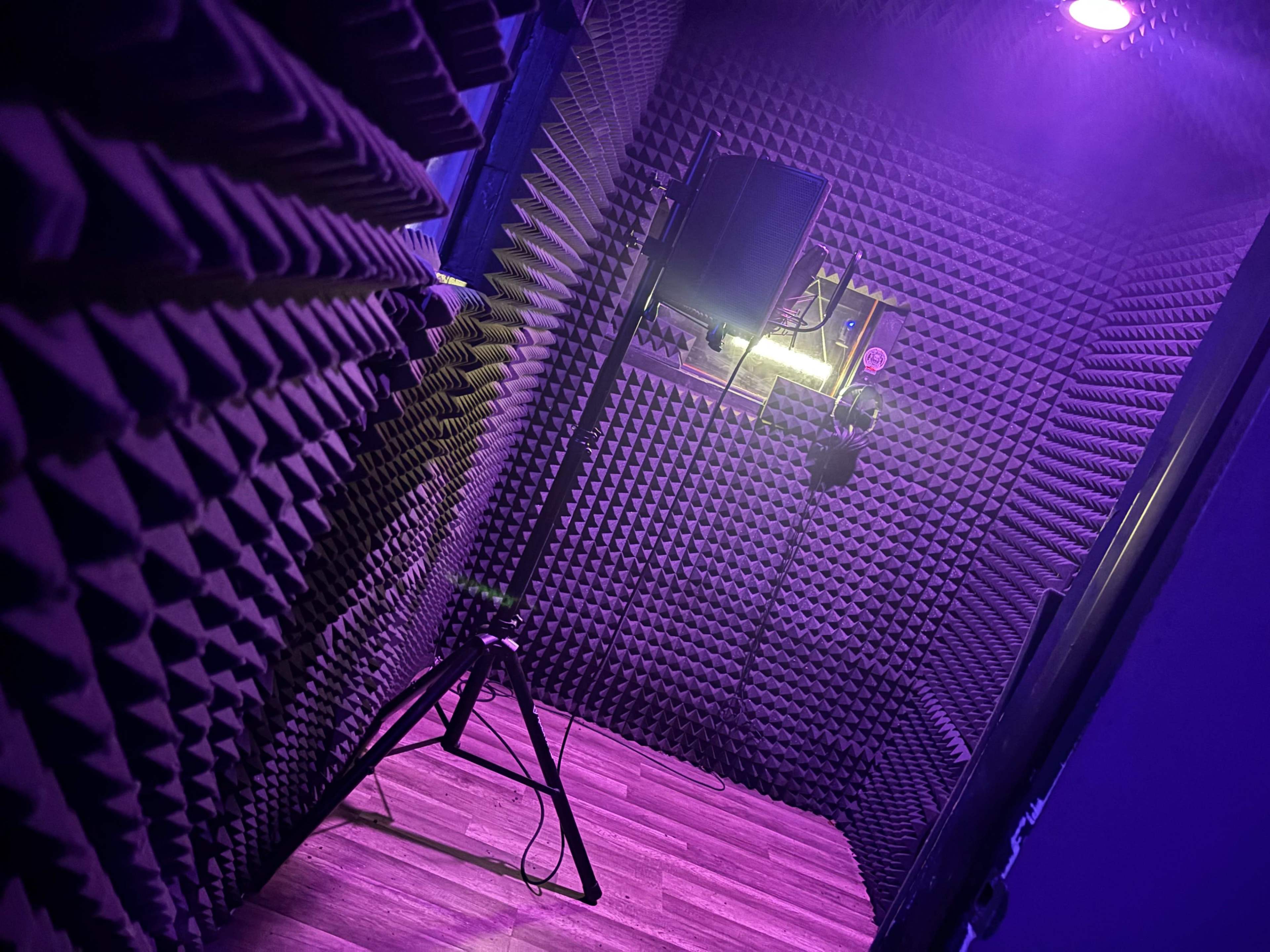 The image shows a soundproof recording studio room with textured acoustic foam on the walls and a tripod in the corner.