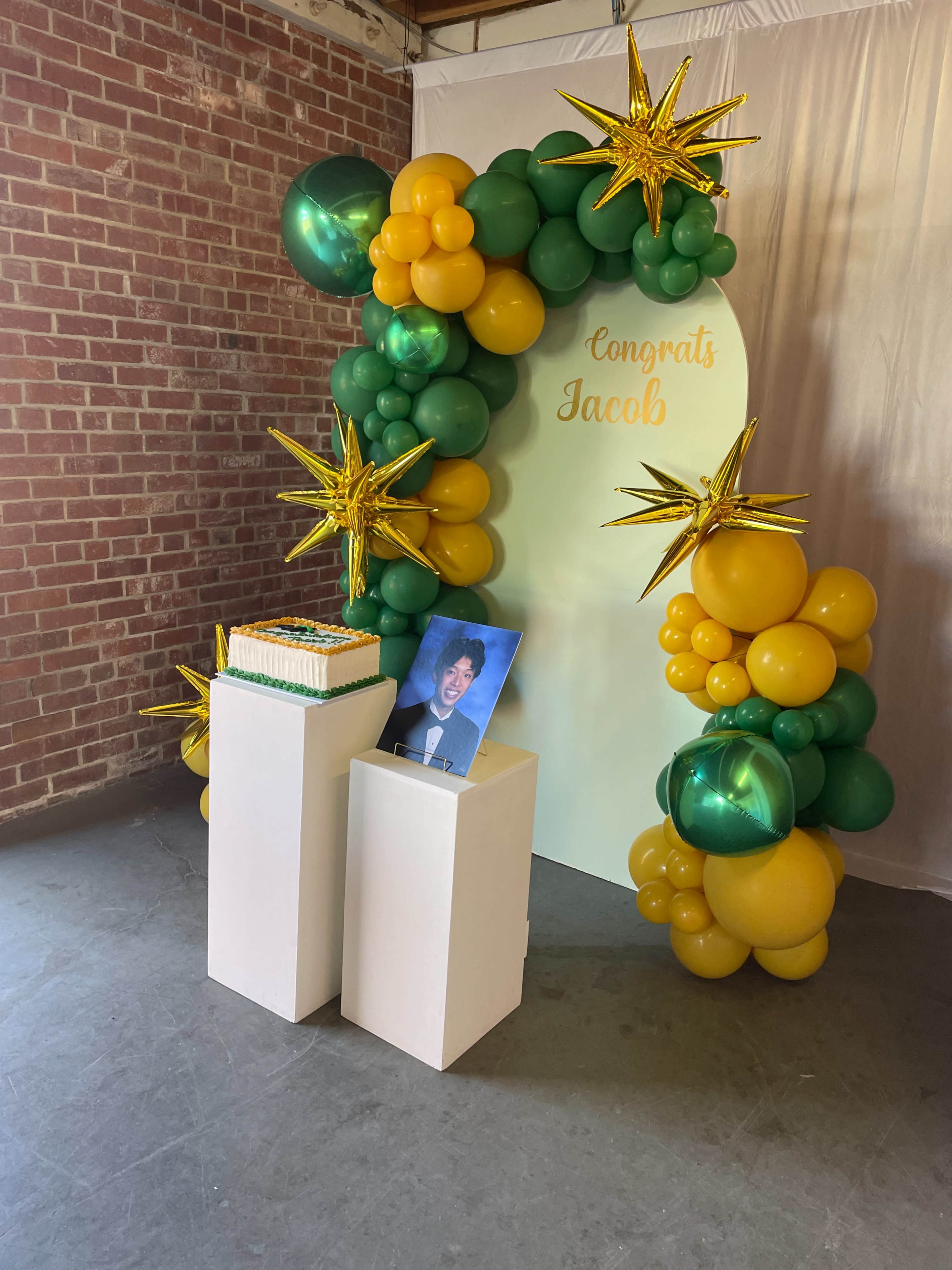 A decorated area featuring a balloon arch in green and gold, a cake on a pedestal, and a framed photo with the text "Congrats Jacob" on the backdrop.