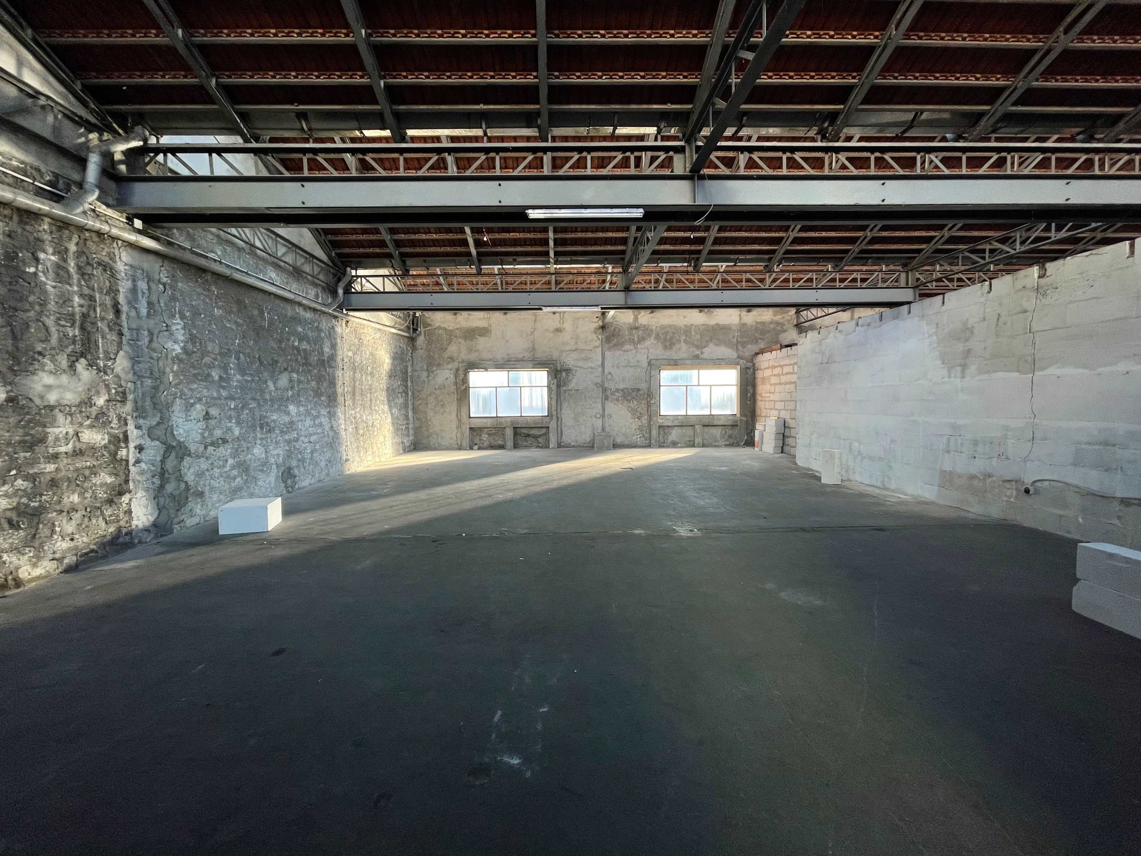 An empty industrial space with exposed brick walls, high ceilings, and two windows allowing natural light to enter.