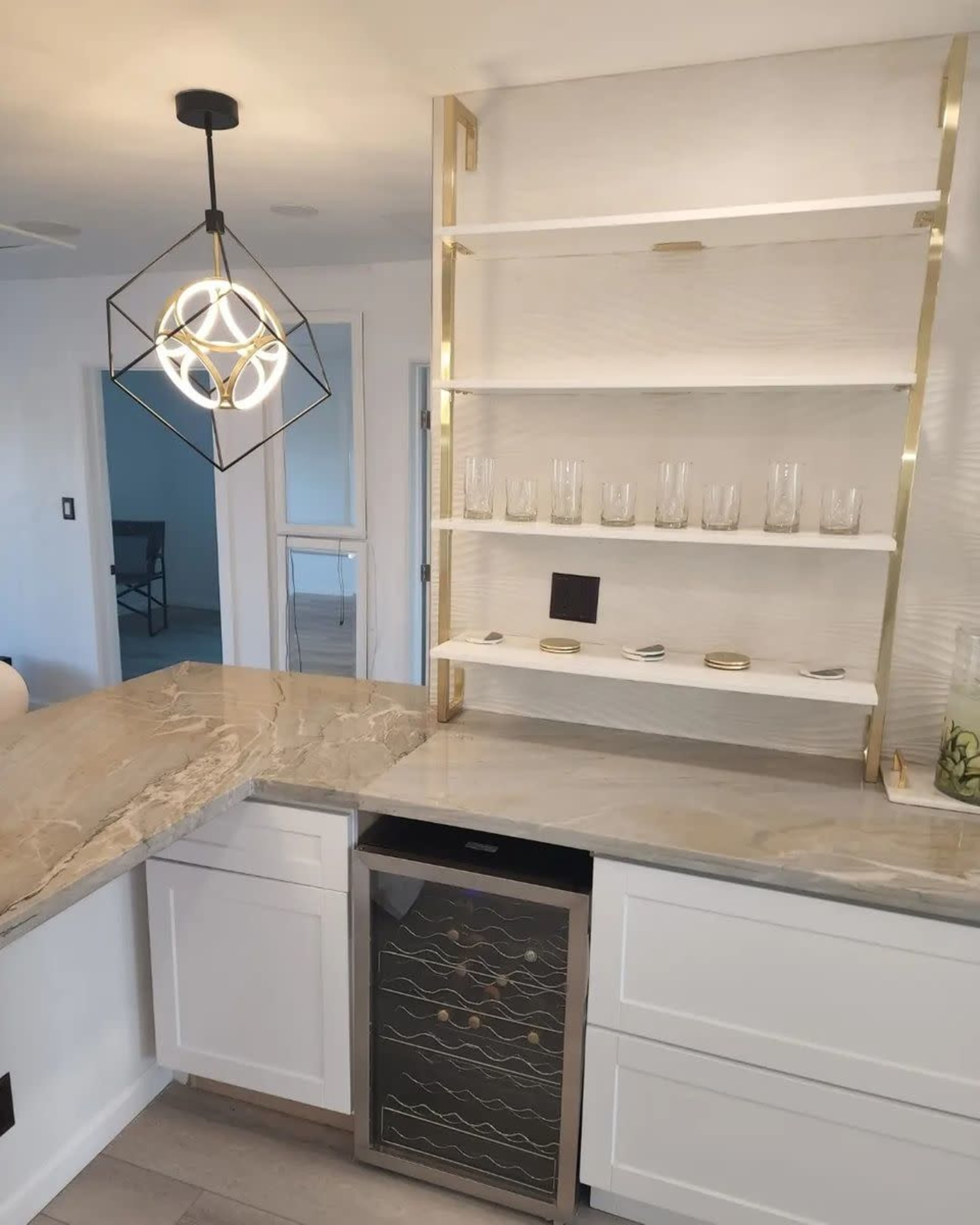A modern kitchen bar area featuring a stone countertop, a gold and black light fixture, and open shelving with glassware above a small refrigerator.