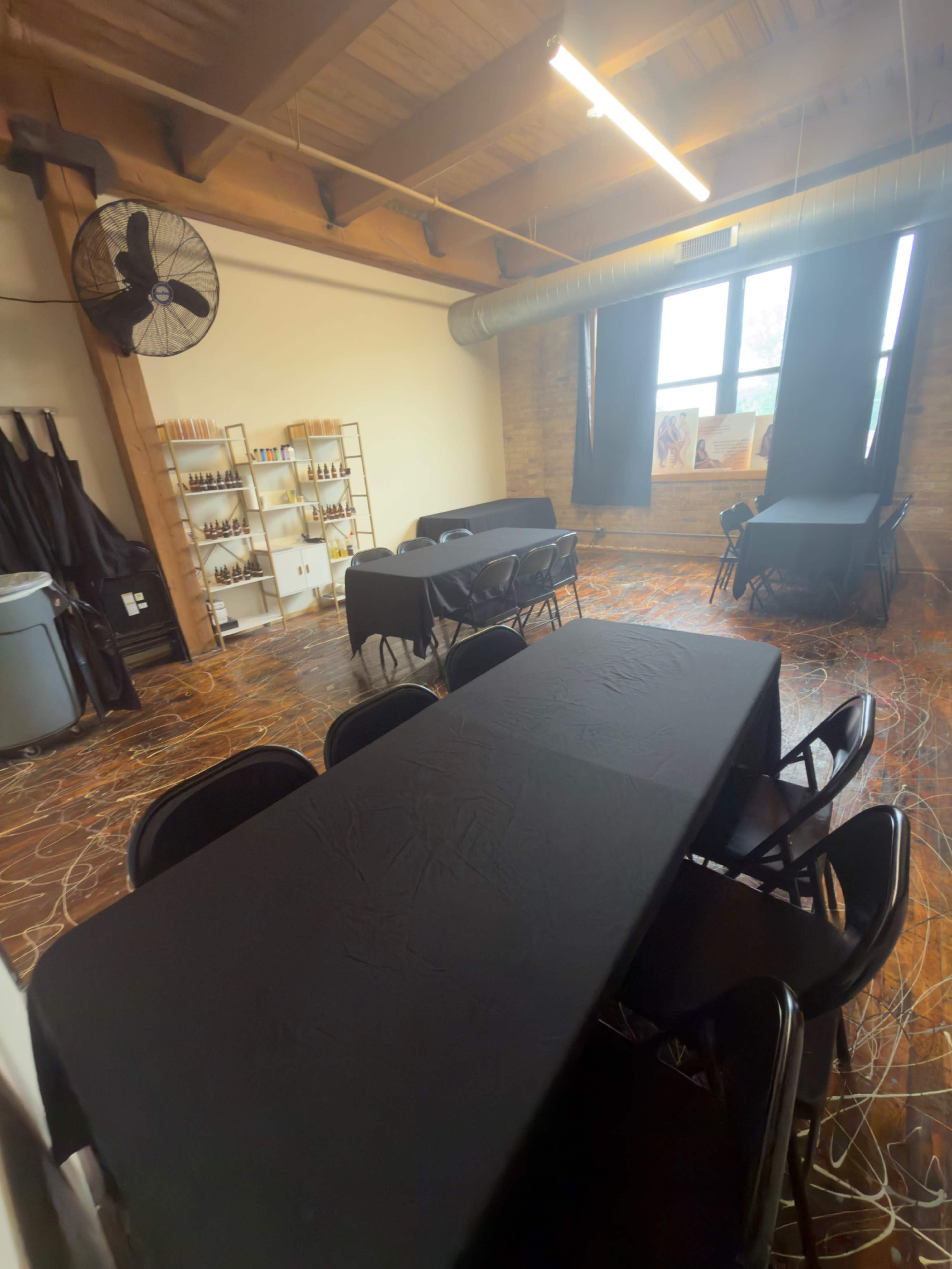 The image shows a spacious room with wooden beams, featuring multiple black tables and chairs arranged, along with shelves displaying bottles against a brick wall.