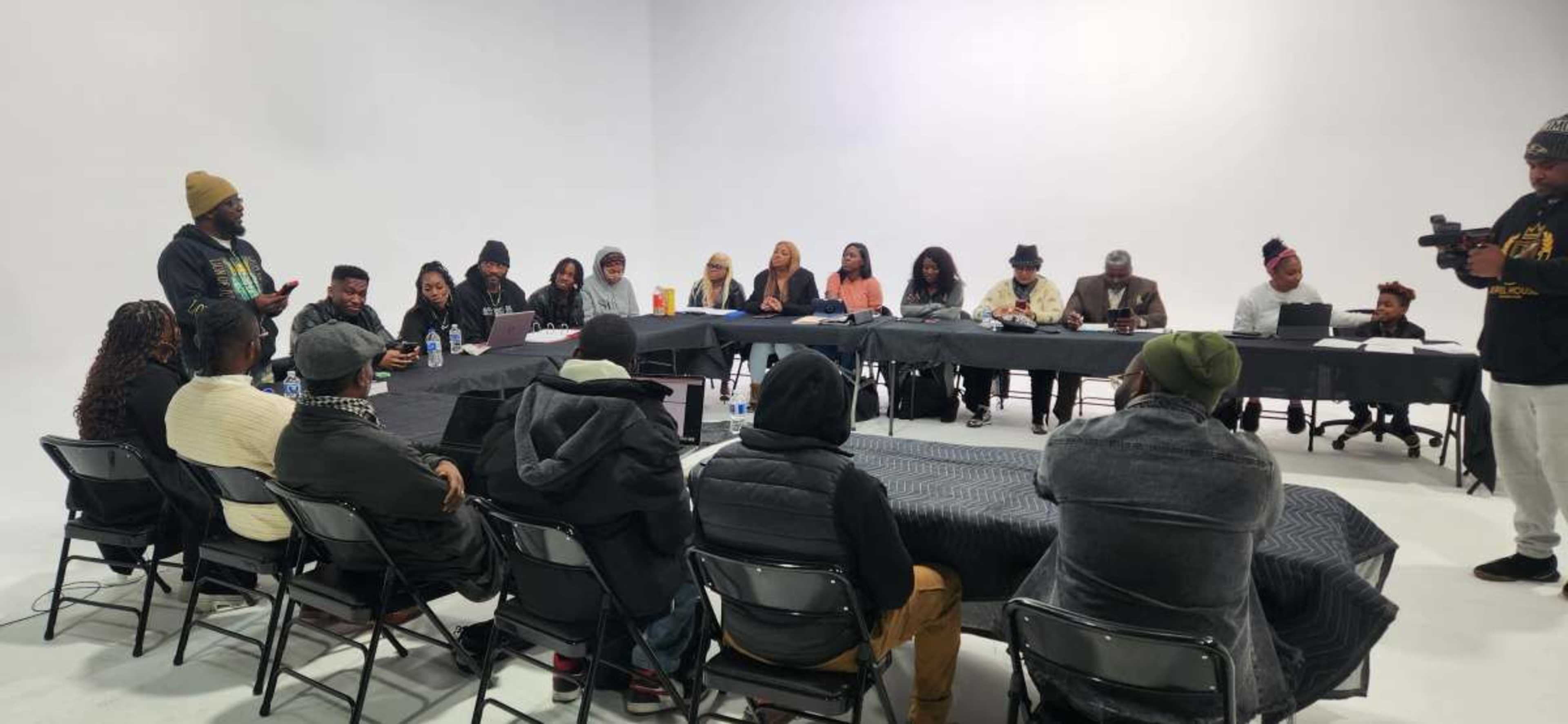 A group of individuals is seated around multiple tables in a spacious, brightly lit meeting room, while one person stands and addresses the assembly.