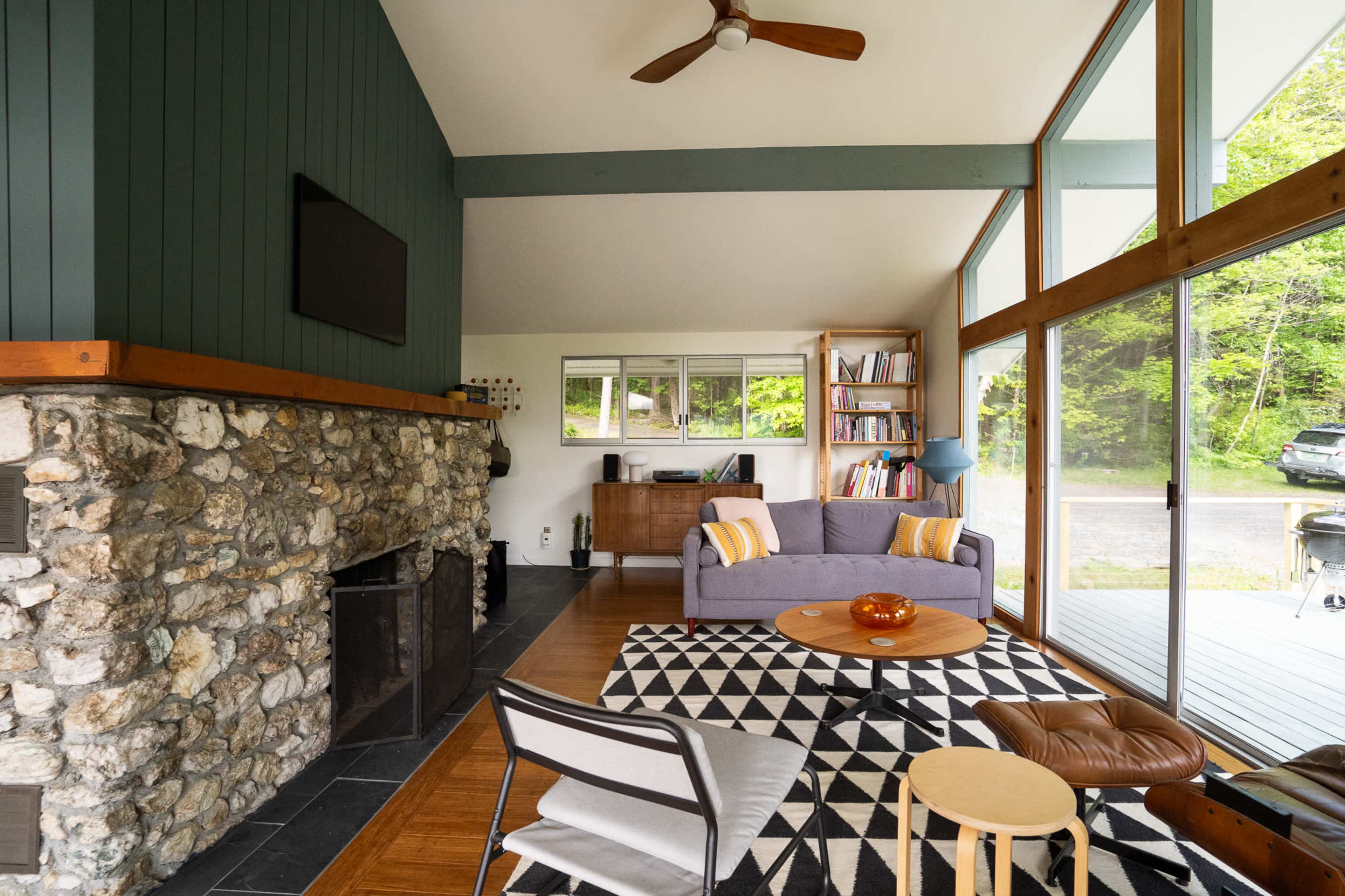 The interior of a modern living room features a stone fireplace, large windows, and a mix of furniture including a gray sofa and a round coffee table on a patterned rug.
