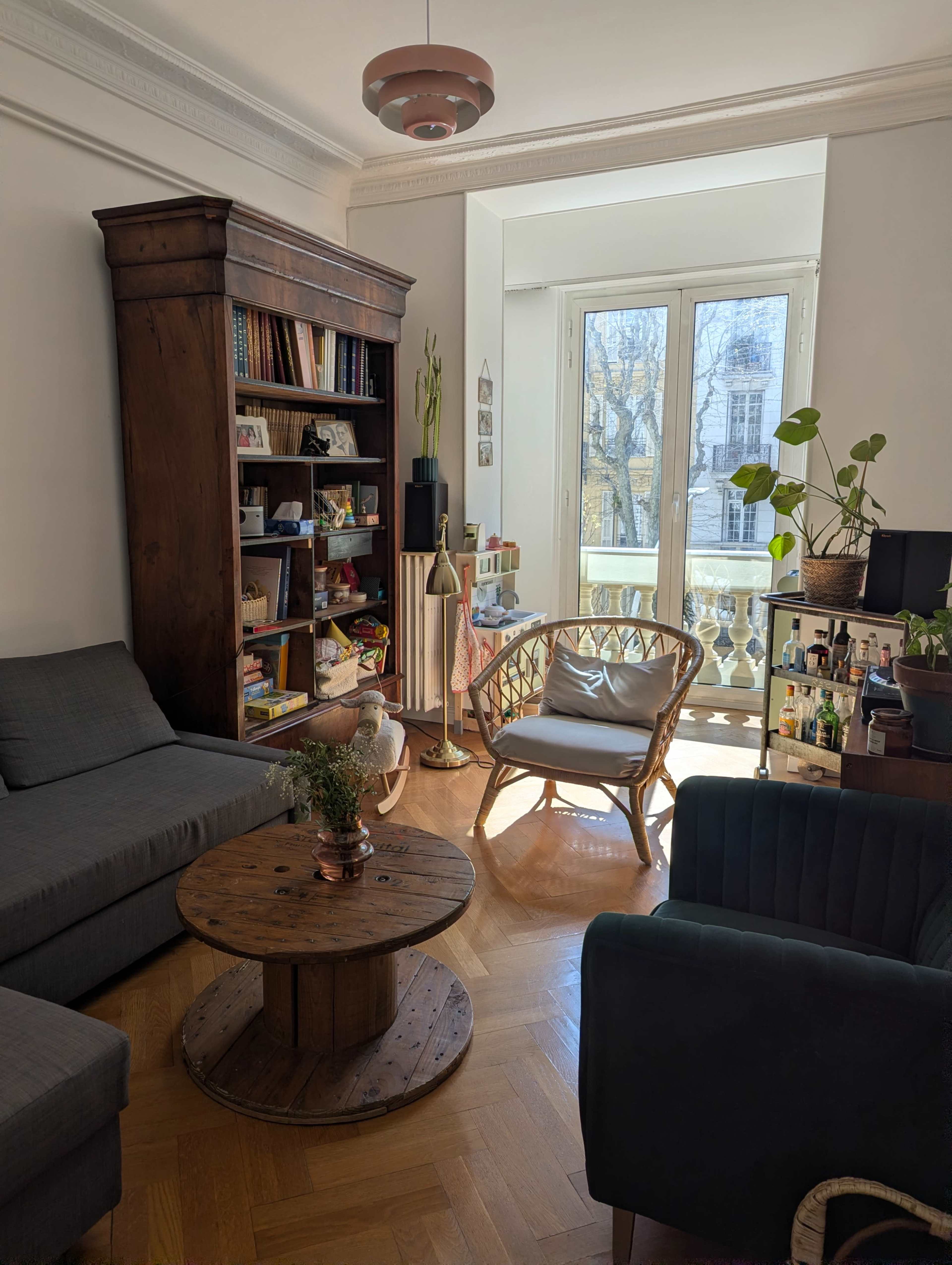 The image shows a cozy living room with a wooden bookshelf filled with books, a round wooden coffee table, a rattan chair, and large windows allowing natural light to enter the space.
