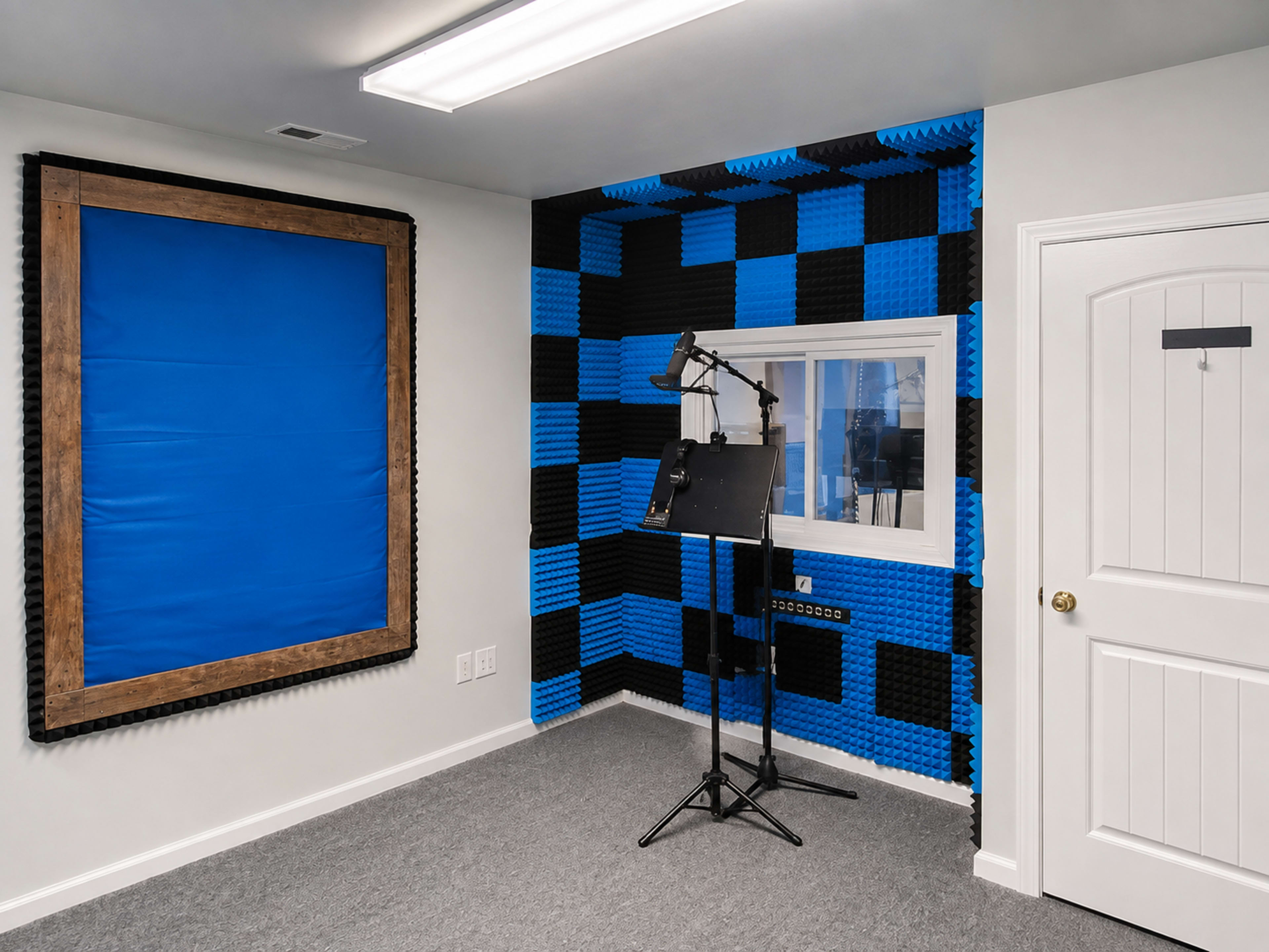 The image shows a soundproof studio with black and blue acoustic foam panels on the walls, a microphone stand, and a window overlooking another room.