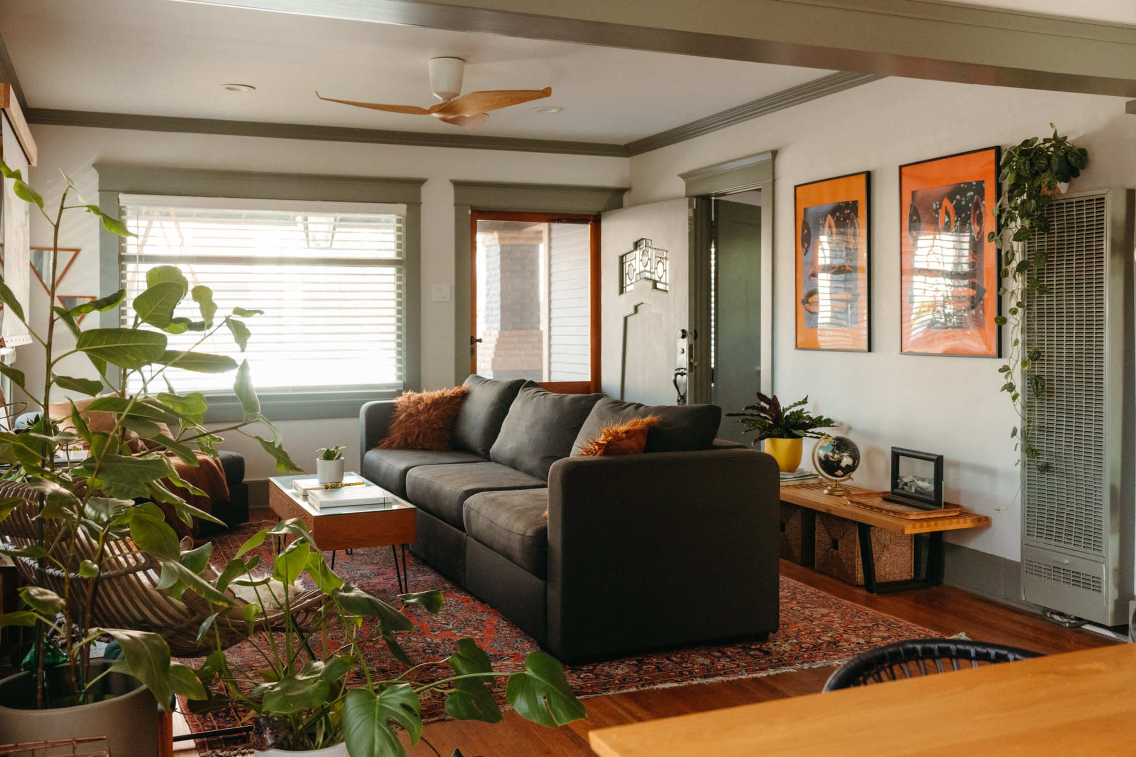 A living room with a gray sofa, potted plants, wooden furniture, and framed artwork on the walls.