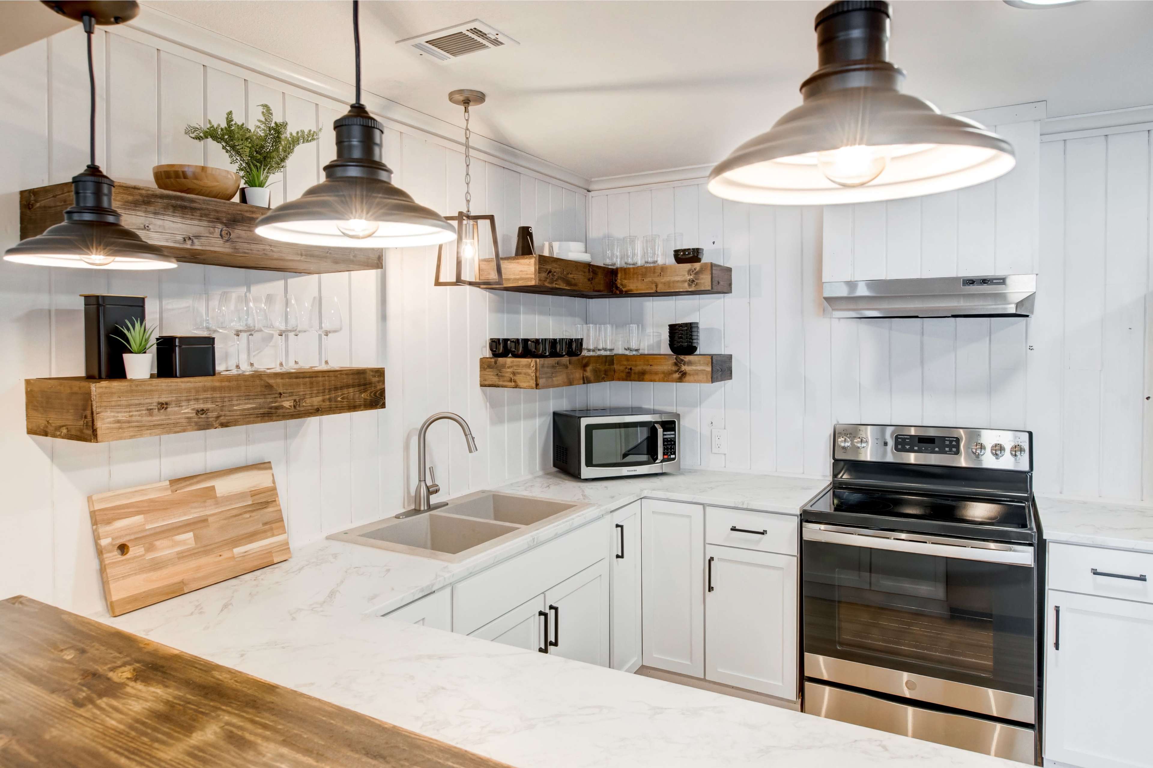 A modern kitchen with white cabinetry, open wooden shelves, a stainless steel stove, a microwave, and a double sink.