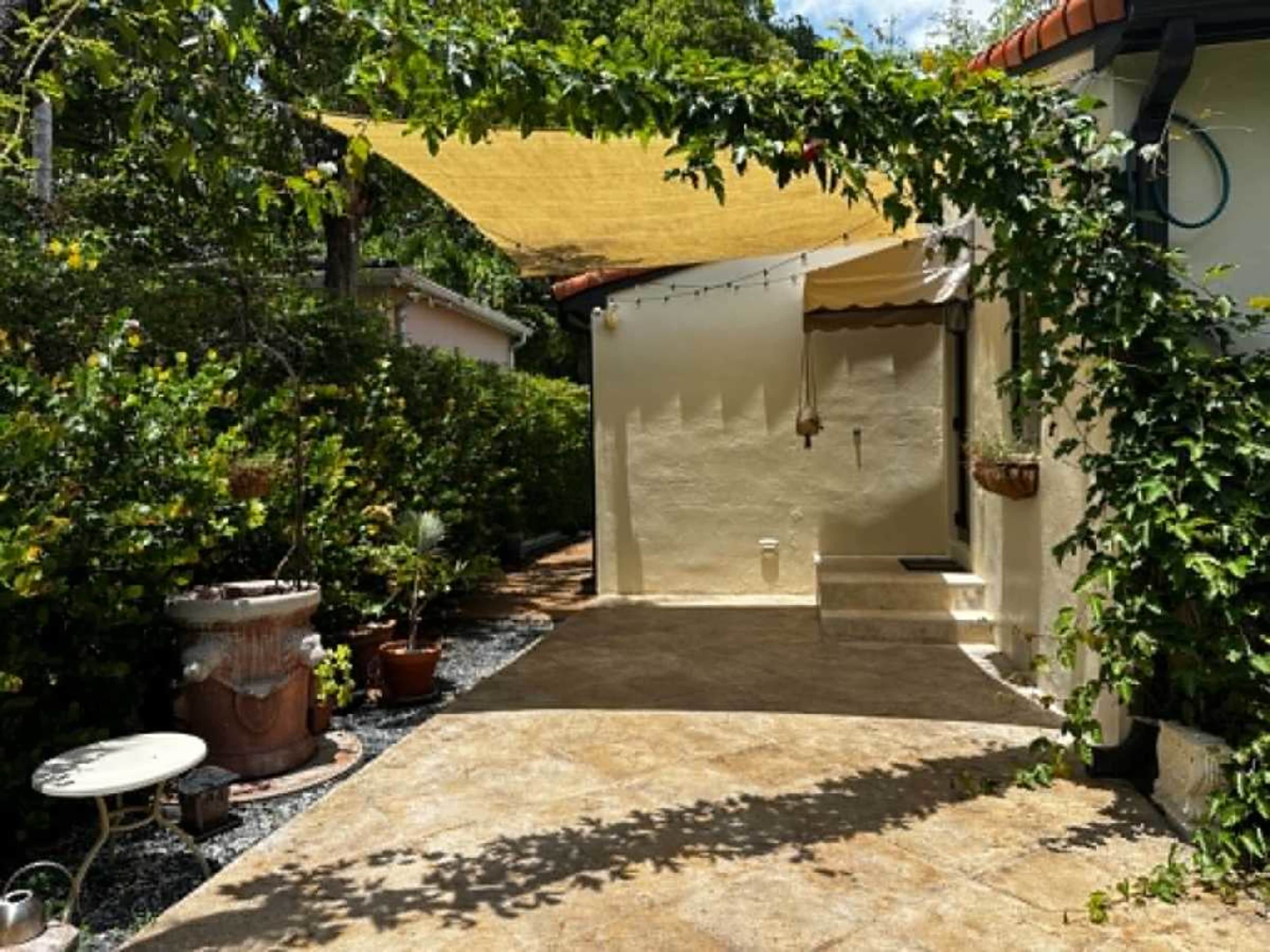 The image shows a patio area surrounded by greenery, with a yellow shade sail overhead and potted plants along the walkway.