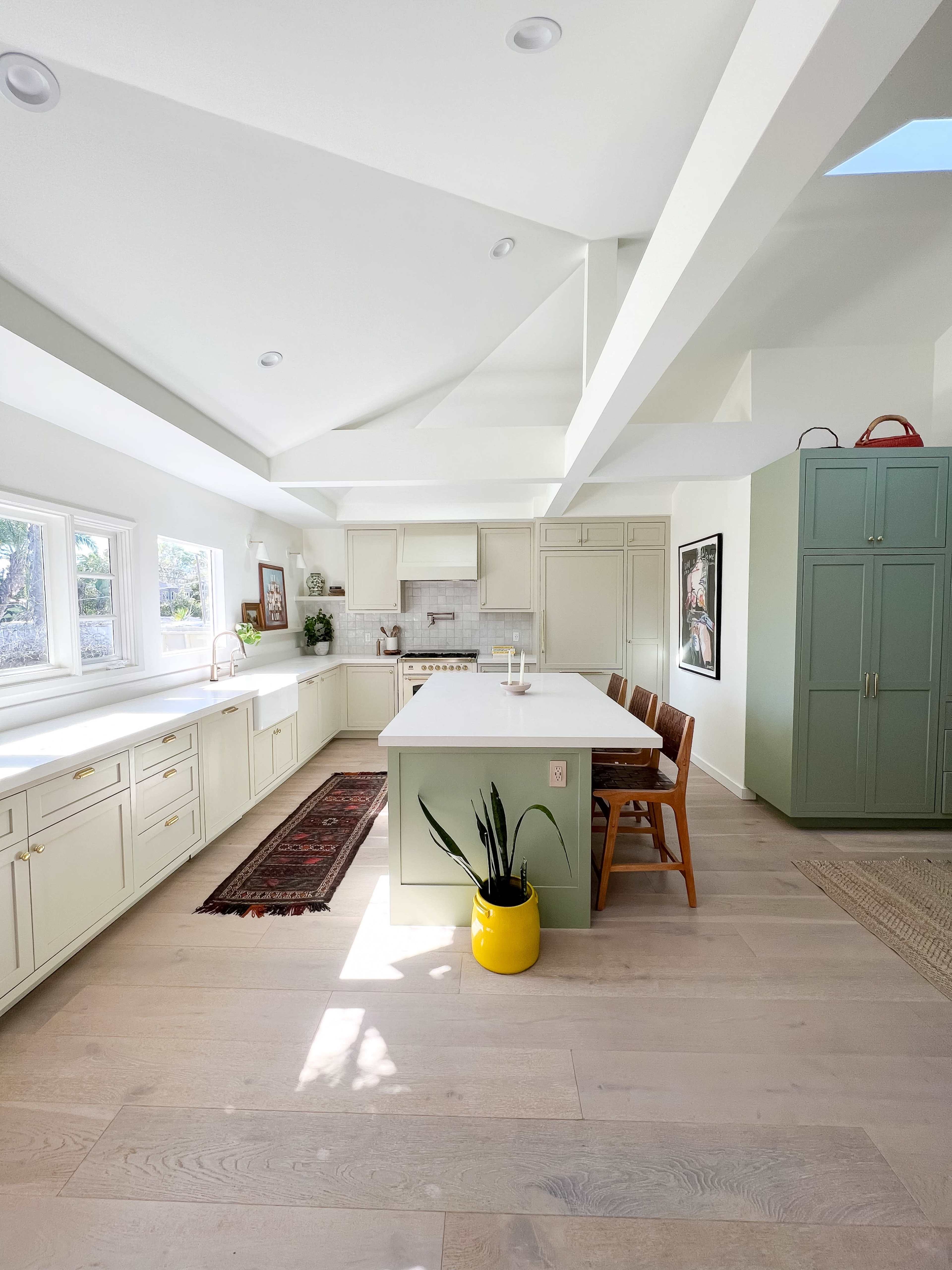 A modern kitchen with a spacious layout, featuring a large central island, white cabinetry, and a green pantry cabinet, along with a colorful rug and a yellow planter.