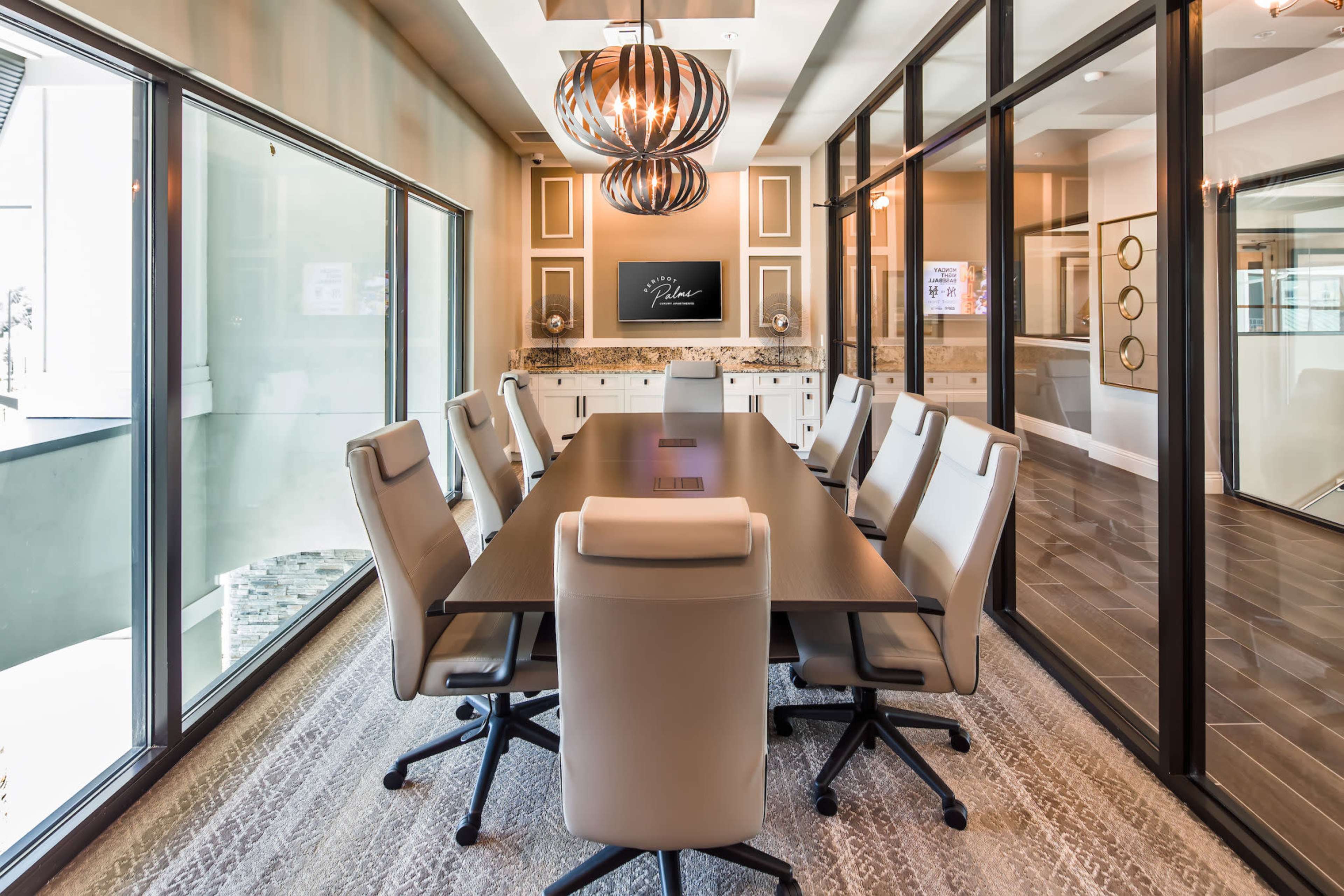 A modern conference room features a large table surrounded by white chairs, with glass walls and a decorative ceiling light.
