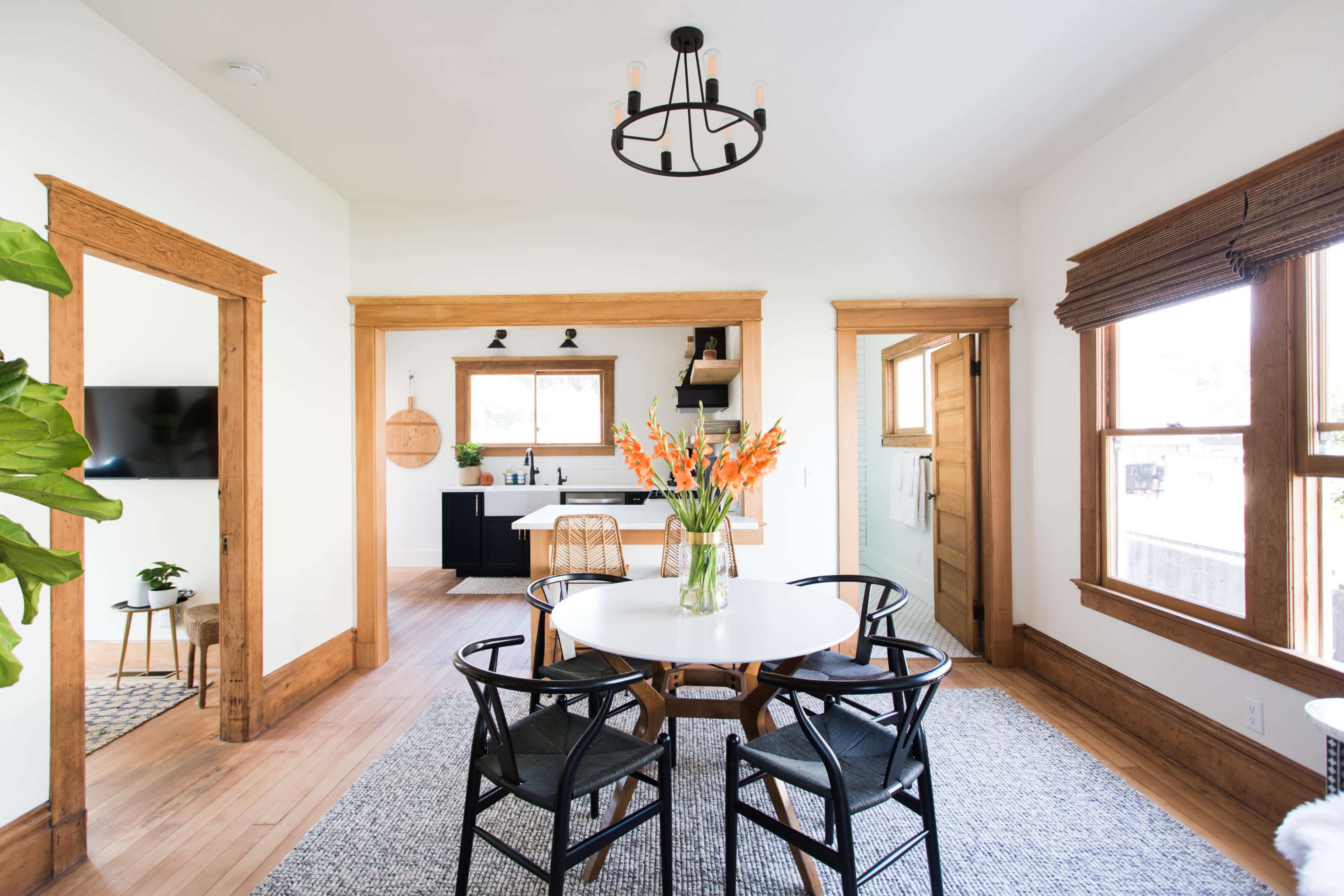 A bright dining area features a round white table surrounded by black chairs, with a view of a modern kitchen and large windows letting in natural light.